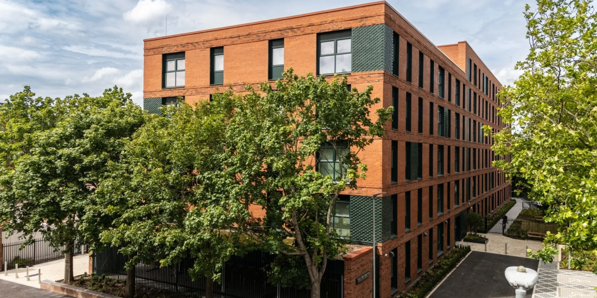 Contemporary red brick residential building with a regular pattern of tall windows and dark textured brick detailing. Mature trees partially screen the façade, with landscaped pathways and a courtyard surrounding the building under a bright, cloudy sky.