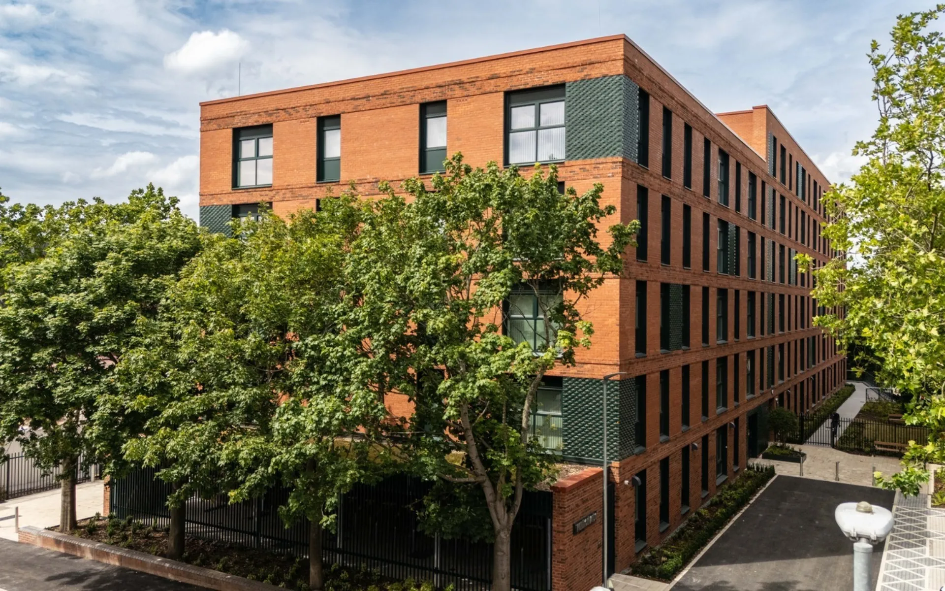 Contemporary red brick residential building with a regular pattern of tall windows and dark textured brick detailing. Mature trees partially screen the façade, with landscaped pathways and a courtyard surrounding the building under a bright, cloudy sky.