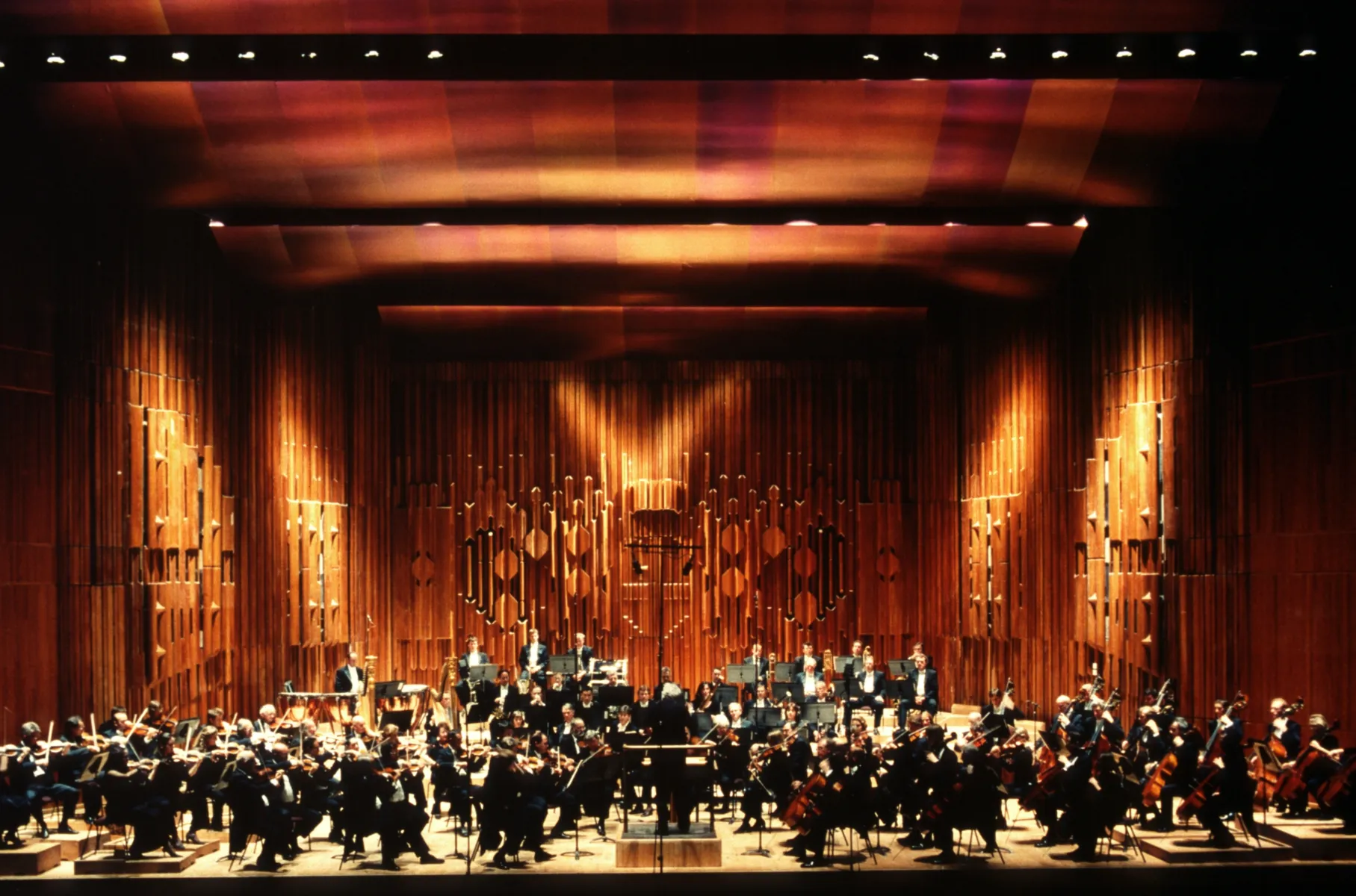 Front view of a large orchestra performing on a wooden stage, with musicians seated around a central conductor, framed by tall sculptural timber acoustic panels and warm overhead lighting.