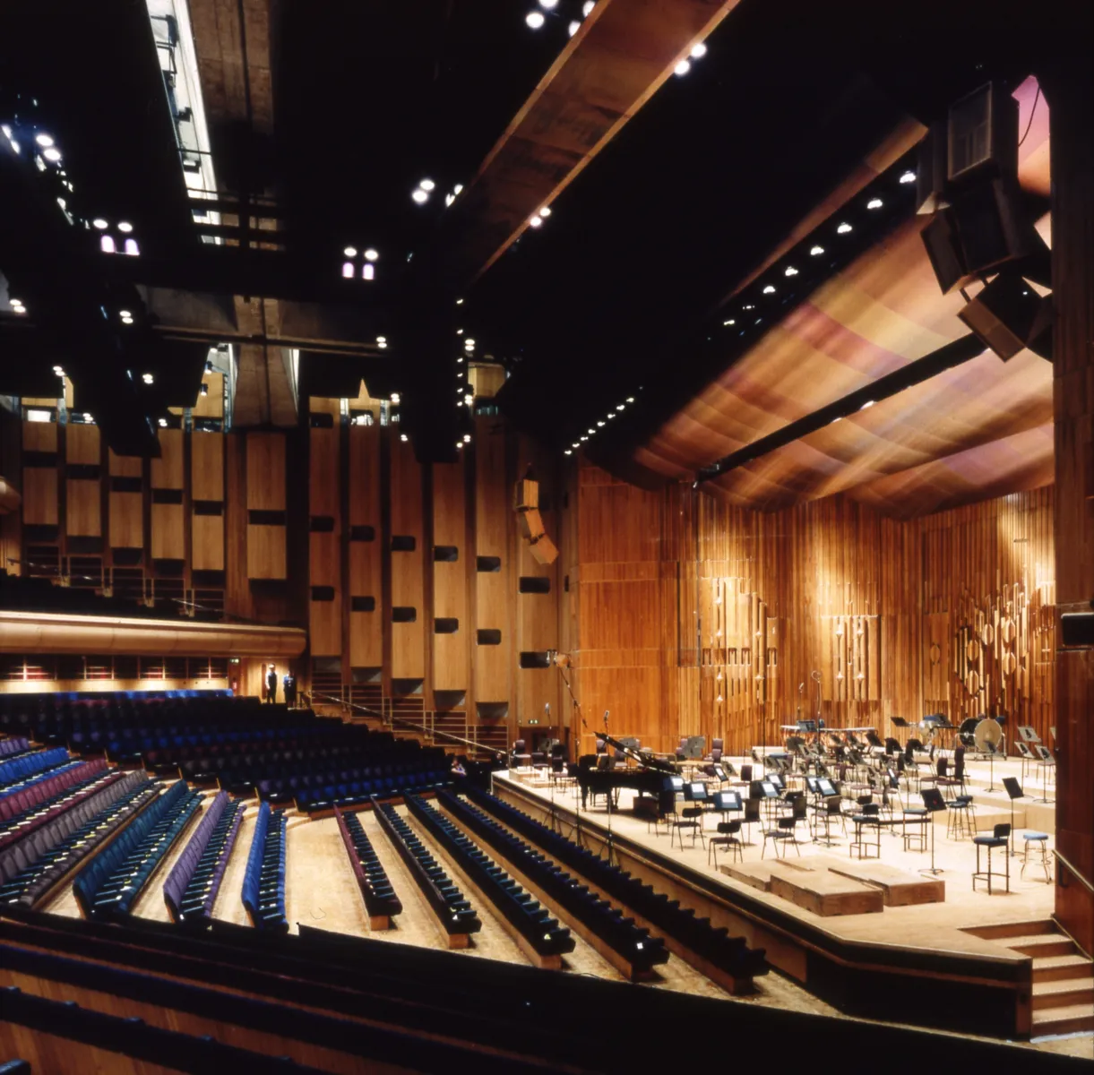 Wide view of a modern concert hall interior with tiered blue seating and a wooden stage set with empty music stands and chairs beneath suspended lighting and acoustic panels.