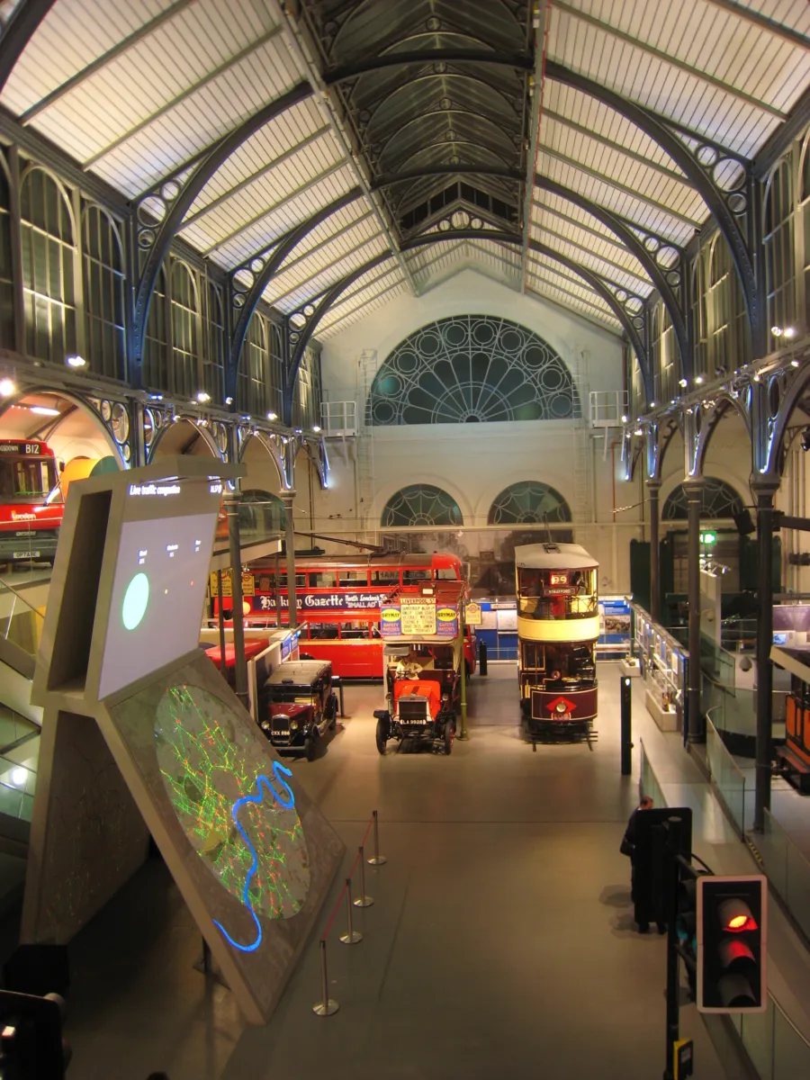 Large museum hall with an arched glass and steel roof, displaying historic buses and vehicles on the gallery floor, with interactive digital screens in the foreground.