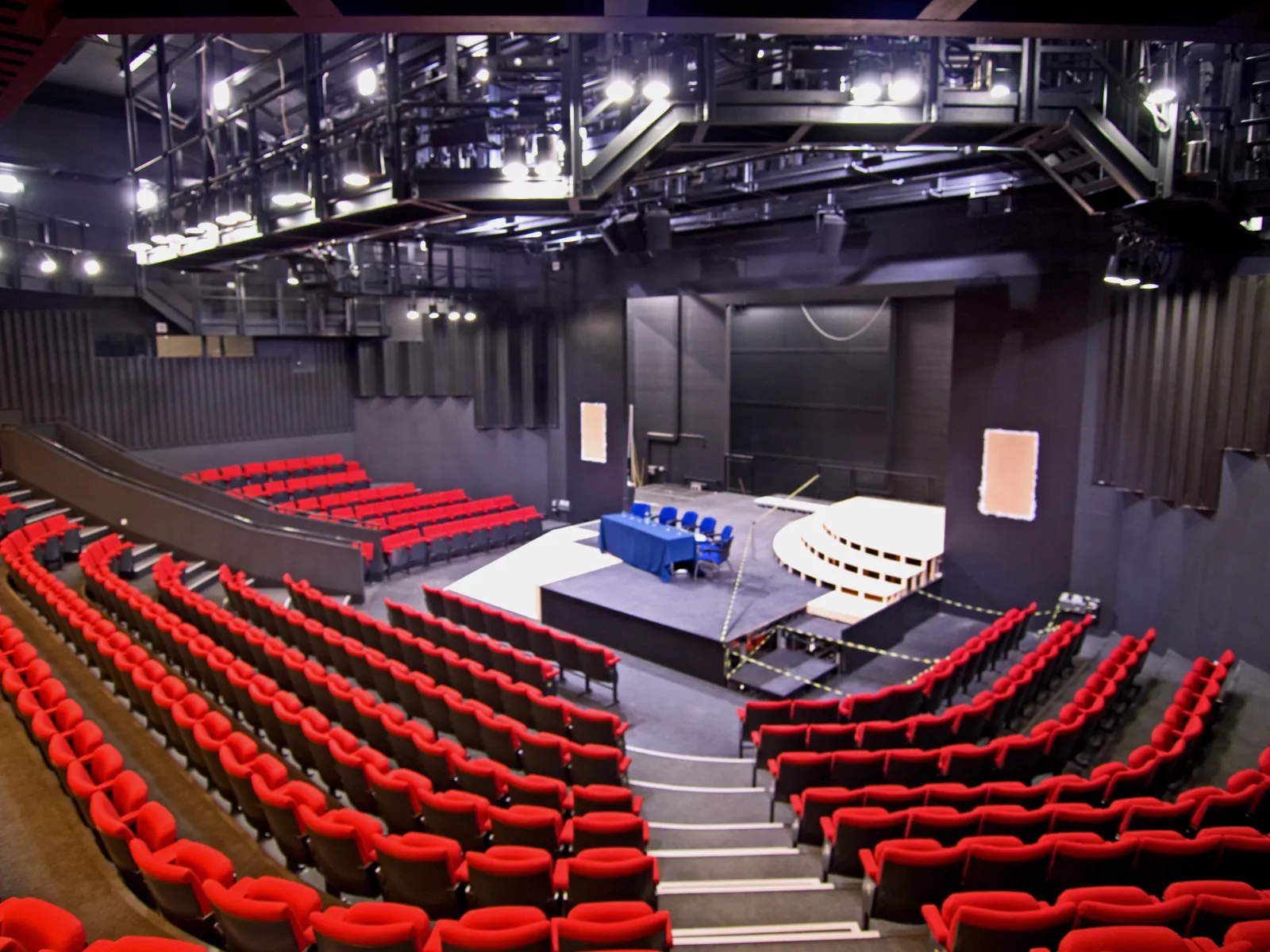 Empty theatre auditorium with curved rows of red seats surrounding a central stage, viewed from the upper seating area under overhead rigging.