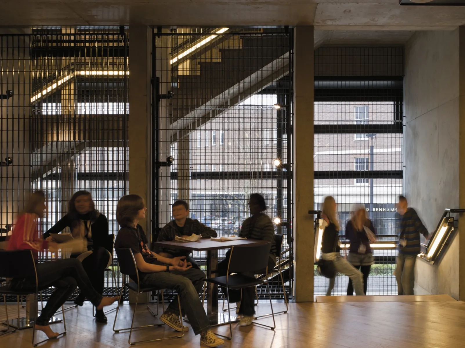 Interior social space with people seated at tables near a stair, framed by concrete columns and metal mesh walls, with daylight visible beyond.