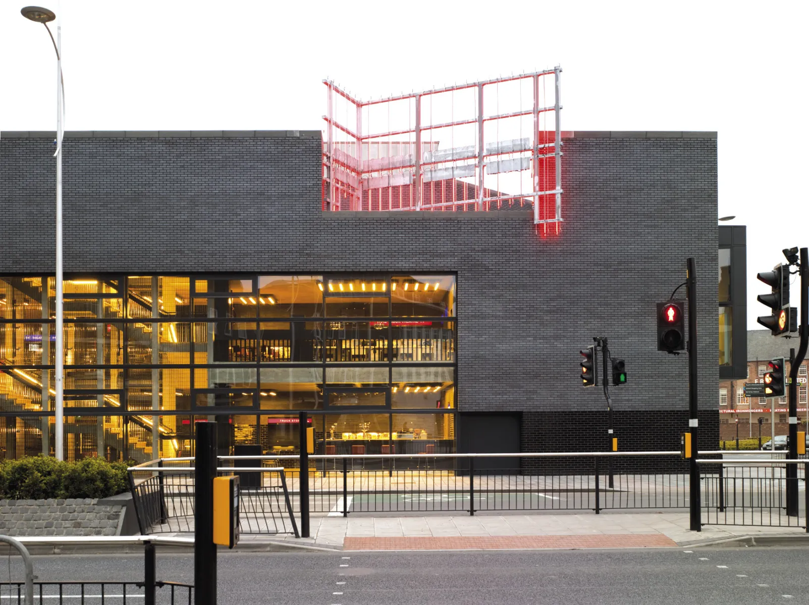 Exterior of a modern brick building at a street junction, with large glowing windows at ground level and a red illuminated steel structure on the roof.