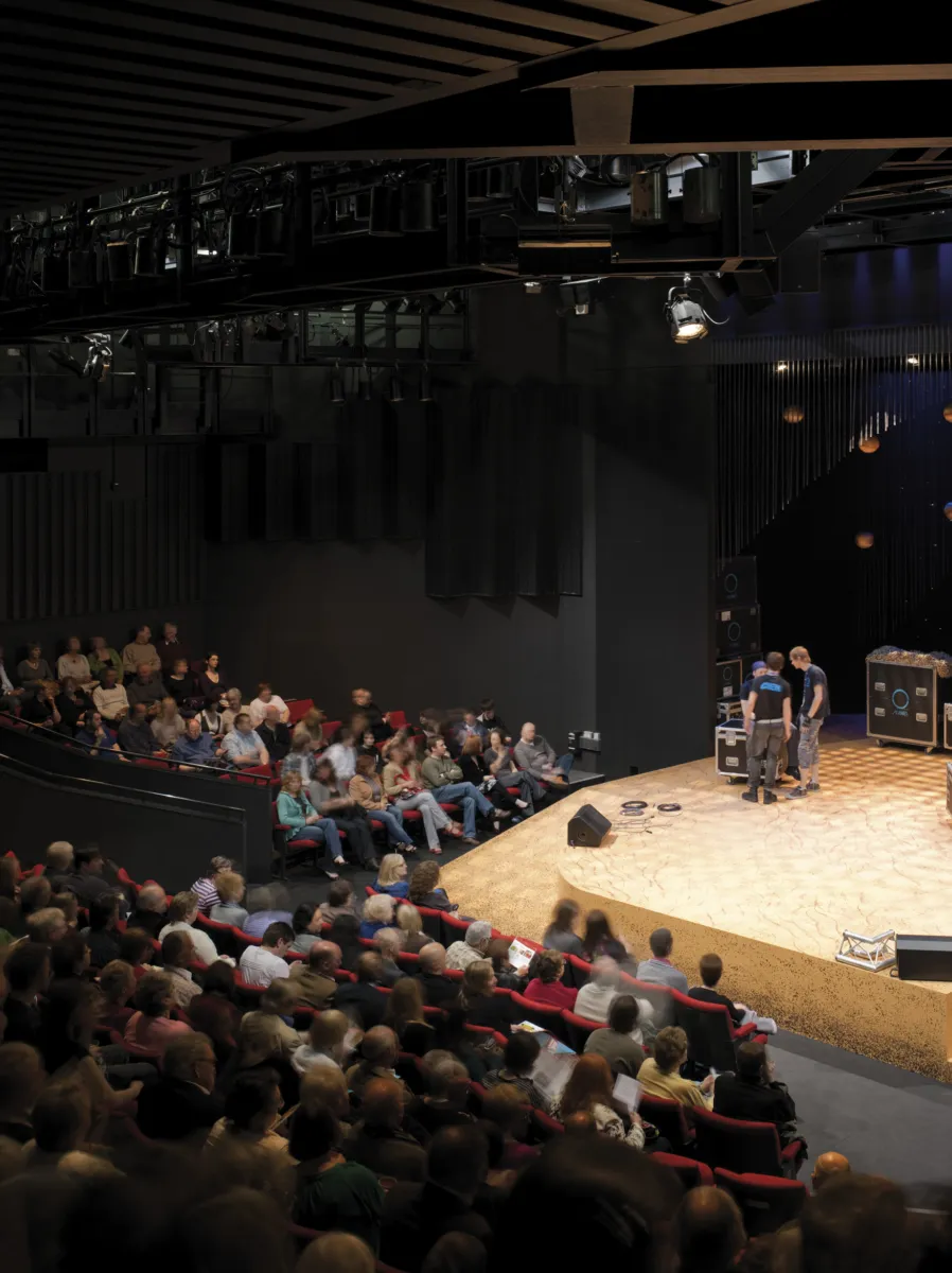 Auditorium interior with a seated audience facing a stage, where equipment and crew are visible under theatrical lighting.