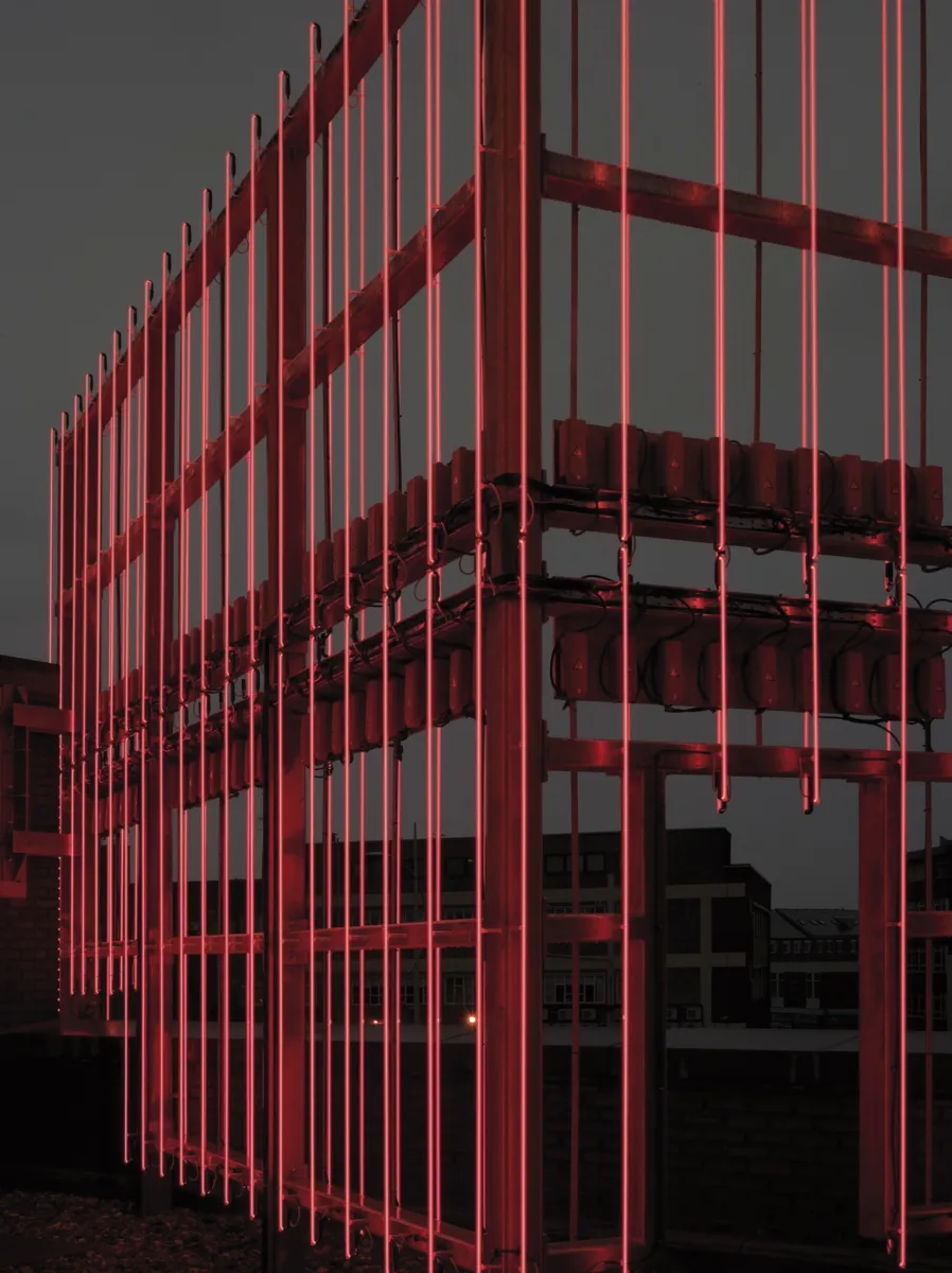 Close-up of a red illuminated steel framework, with vertical glowing light rods mounted to a metal structure against a dark sky.