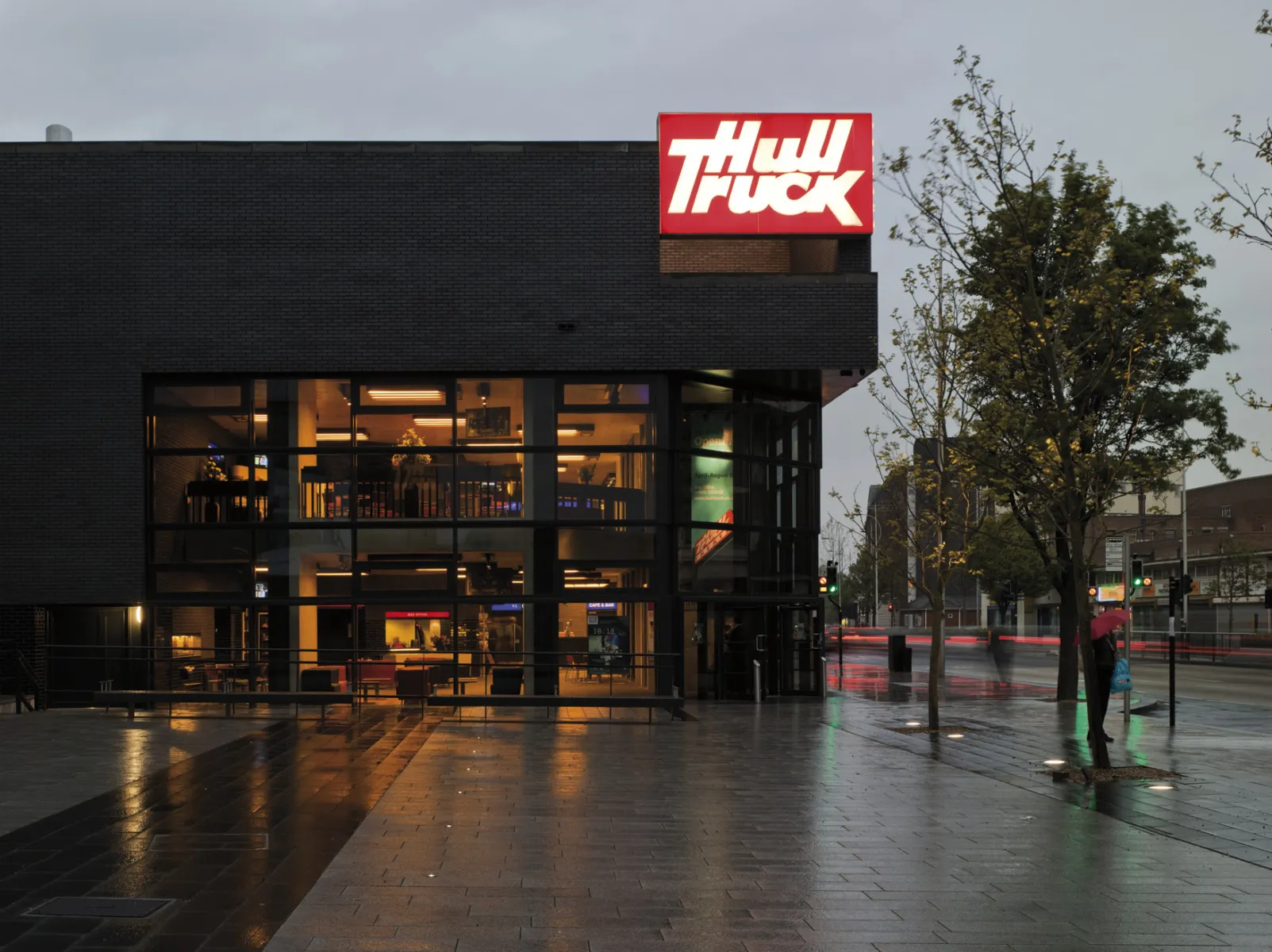 Evening exterior of a theatre building with lit windows and a bright red illuminated sign, reflected on wet paving after rain.