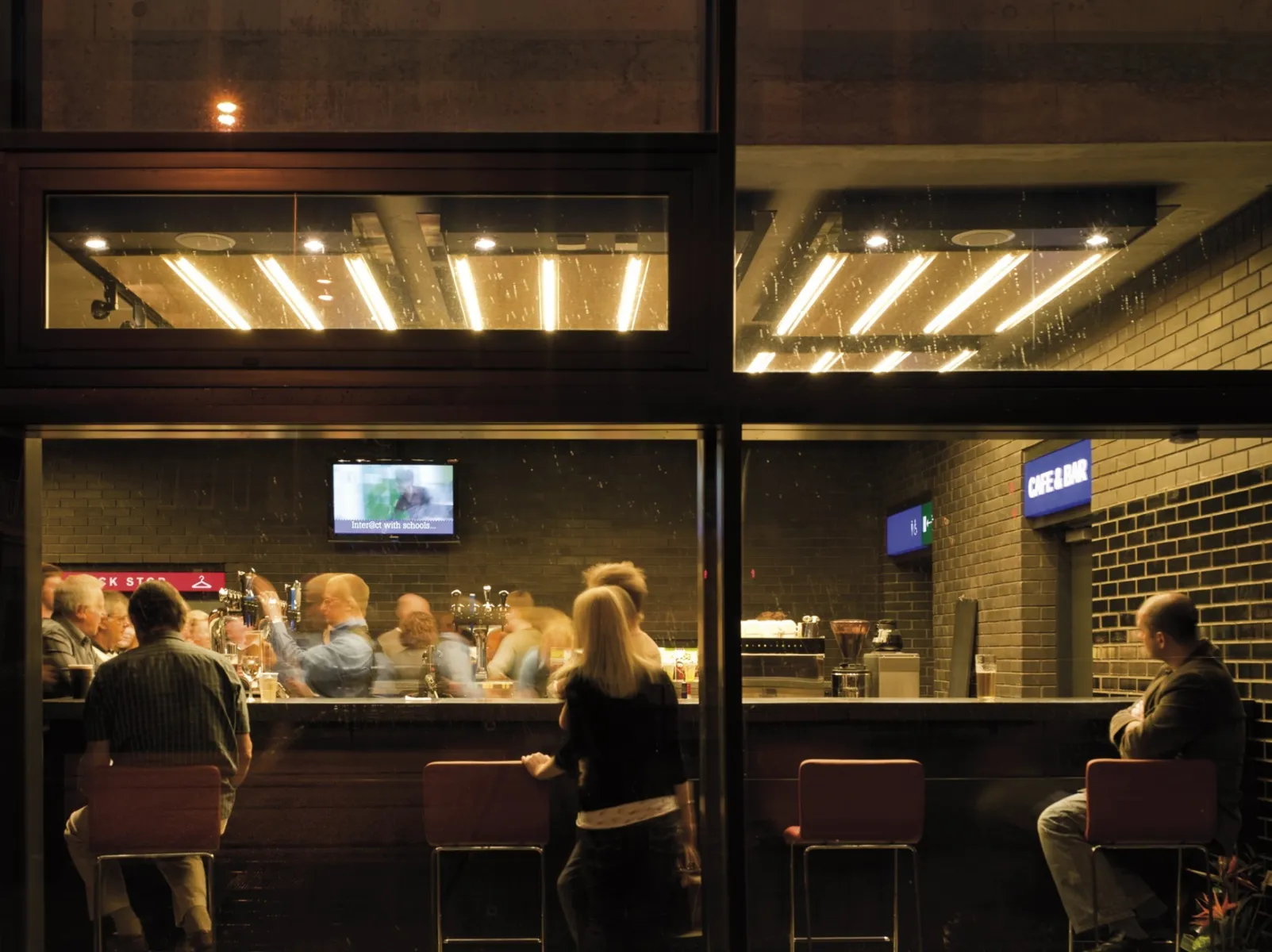 View through a large glass window into a bar area, where people sit and stand under warm lighting, with bar stools lined along the glass.