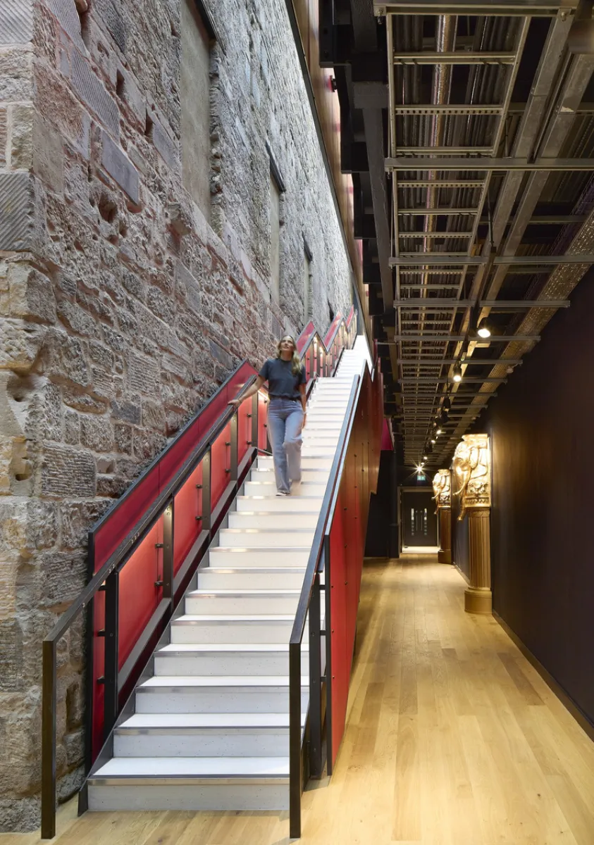 Images shows a corridor with a red staicase, set next to an old stone wall