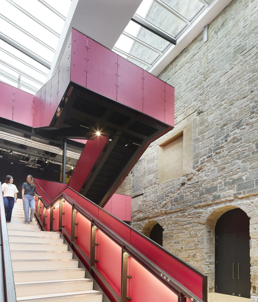 image shows a pink staircase with skylights in the roof, and an old stone wall