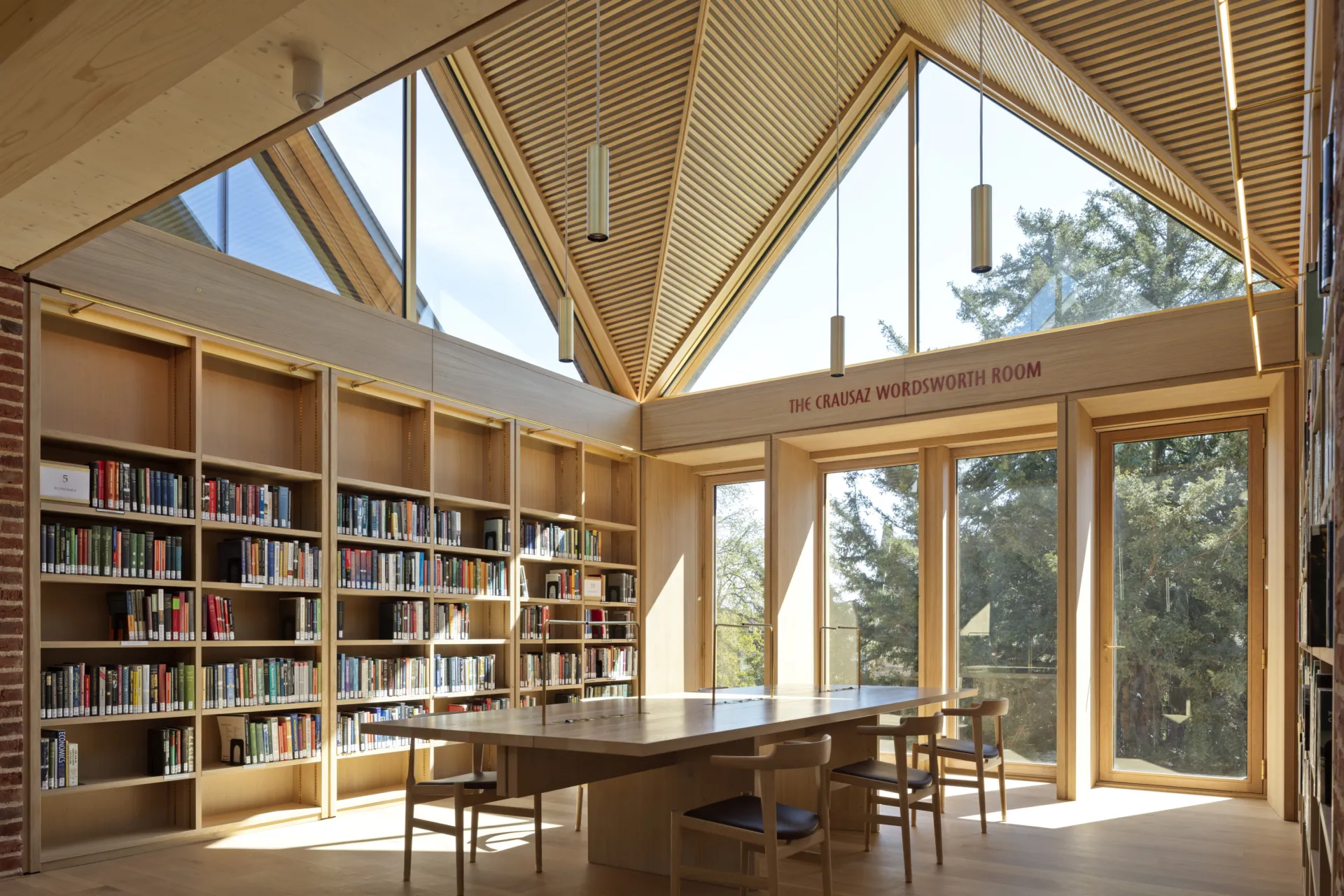A light-filled library with light-coloured wood. Bookshelves line the walls and a large desk surrounded by chairs sits in the middle of the room Daylight pours in through the large windows.