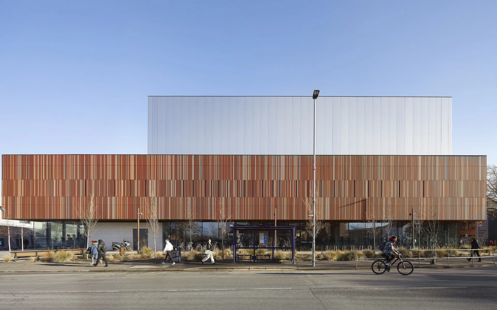 Exterior of the sports centre of a clear blue-sky day. The ground floor of the building is glazed with a large upper section that appears to be clad in wood.