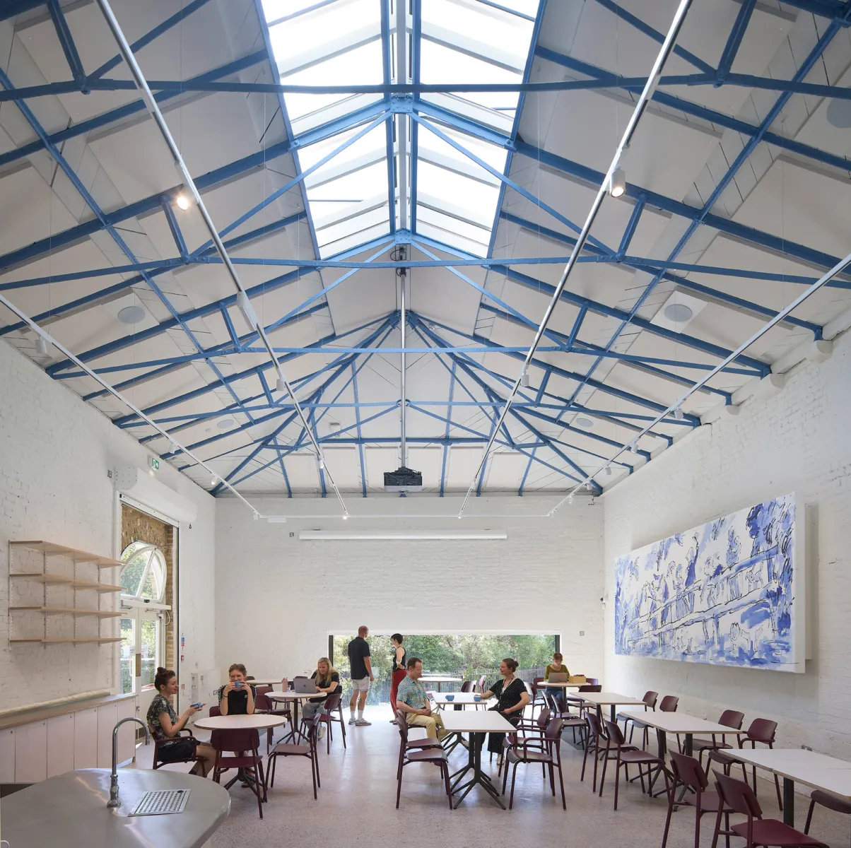 Bright café space with white-painted brick walls and blue steel roof trusses, where café tables and chairs are arranged across the room and several people sit, talk, and use laptops beneath a large glazed opening to greenery outside.