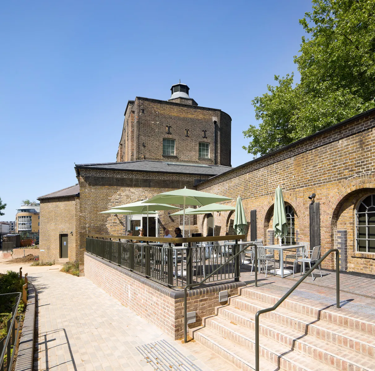 Outdoor terrace with tables and pale green umbrellas set against historic brick buildings, where people sit at tables on a raised platform accessed by steps, with trees and blue sky in the background.