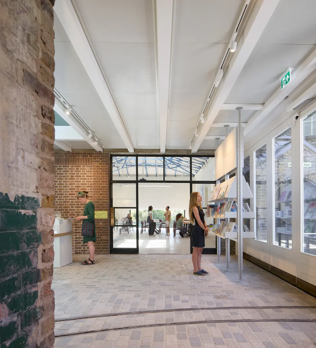 Interior view of a brick-lined entrance space with a cobbled floor and white ceiling beams, where display shelving lines the right-hand wall; several people browse books while others are visible through glazed doors in a café area beyond.