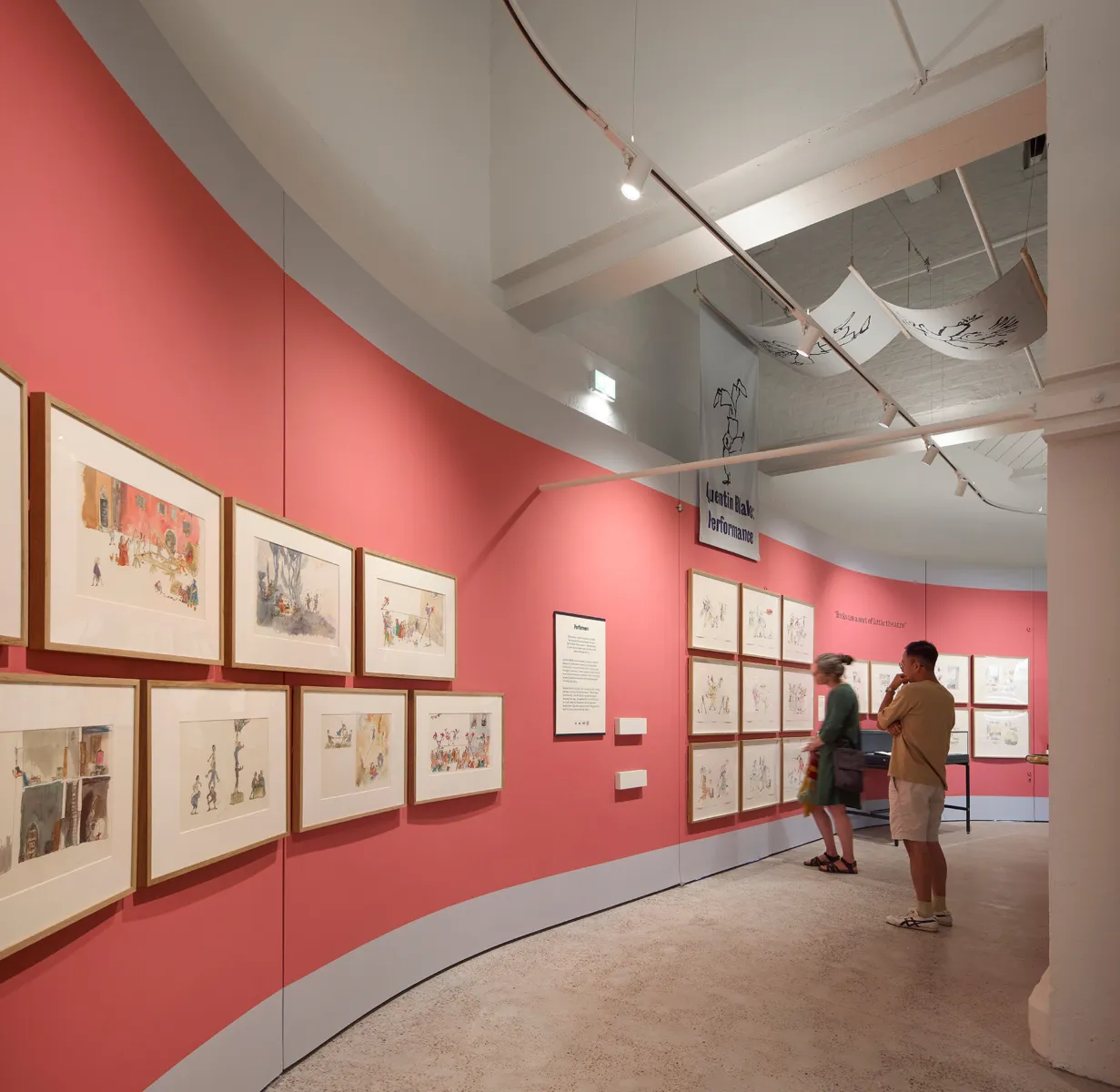 Curved gallery with pink walls displaying framed illustrations, with suspended fabric banners above; two visitors stand viewing the artwork along the wall under track lighting.