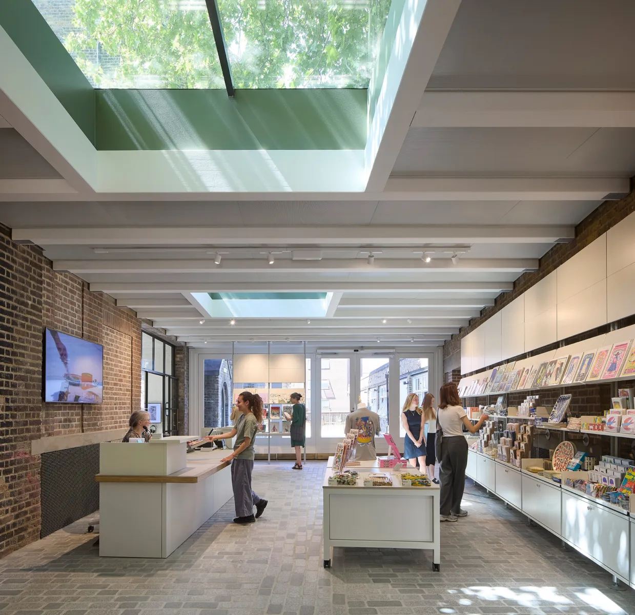 Interior shop with exposed brick walls, white joinery and a skylight above, where a counter and central display tables present books and merchandise; several people browse and make a purchase.