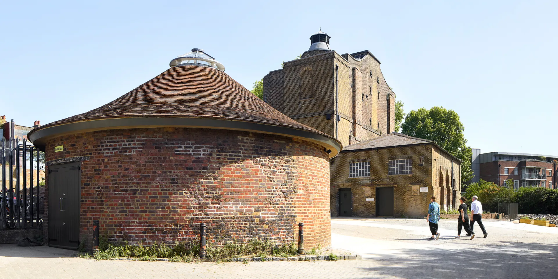 Circular brick building with a conical tiled roof in the foreground set within an open paved courtyard. Behind it stands a larger historic brick structure with a square tower and small windows. Three people walk across the courtyard.