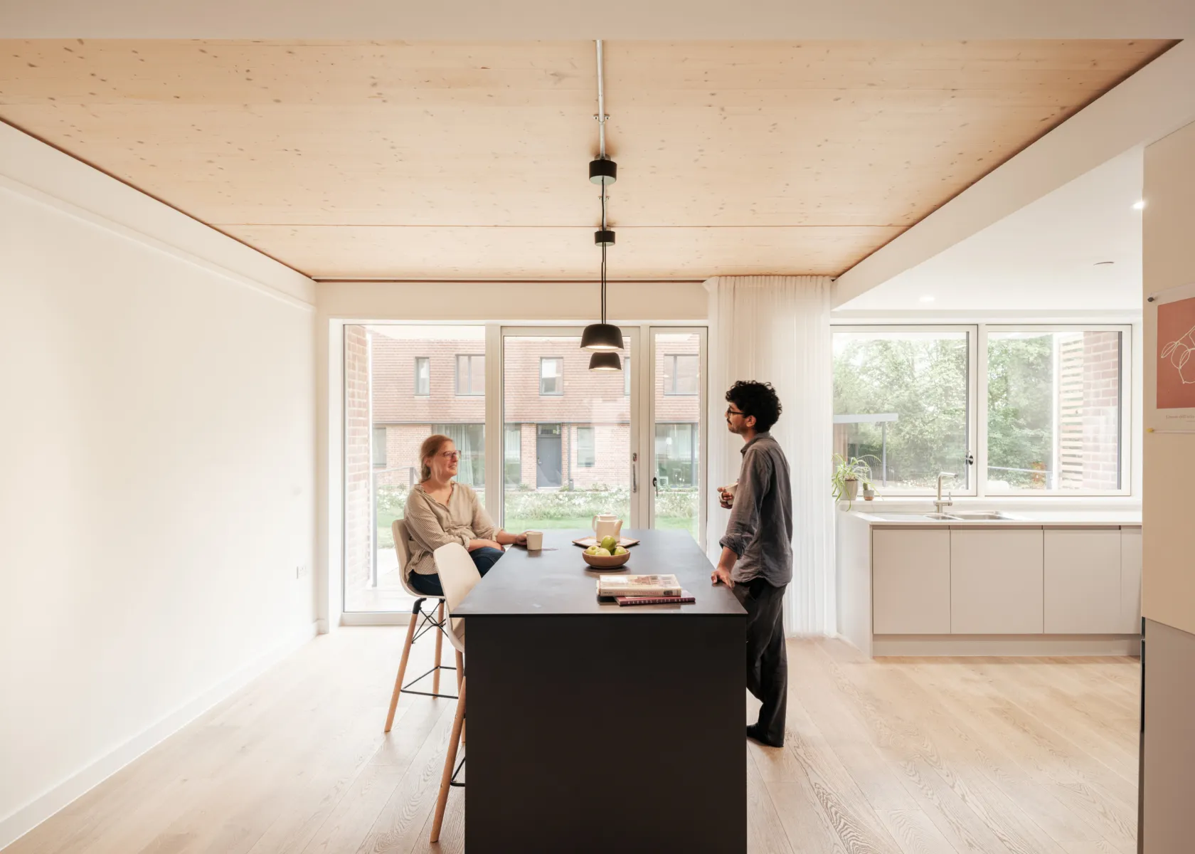 Bright, open-plan kitchen and dining space with a timber ceiling and large windows. Two people stand and sit at a central kitchen island, talking, with daylight overlooking a brick courtyard and modern residential buildings beyond.
