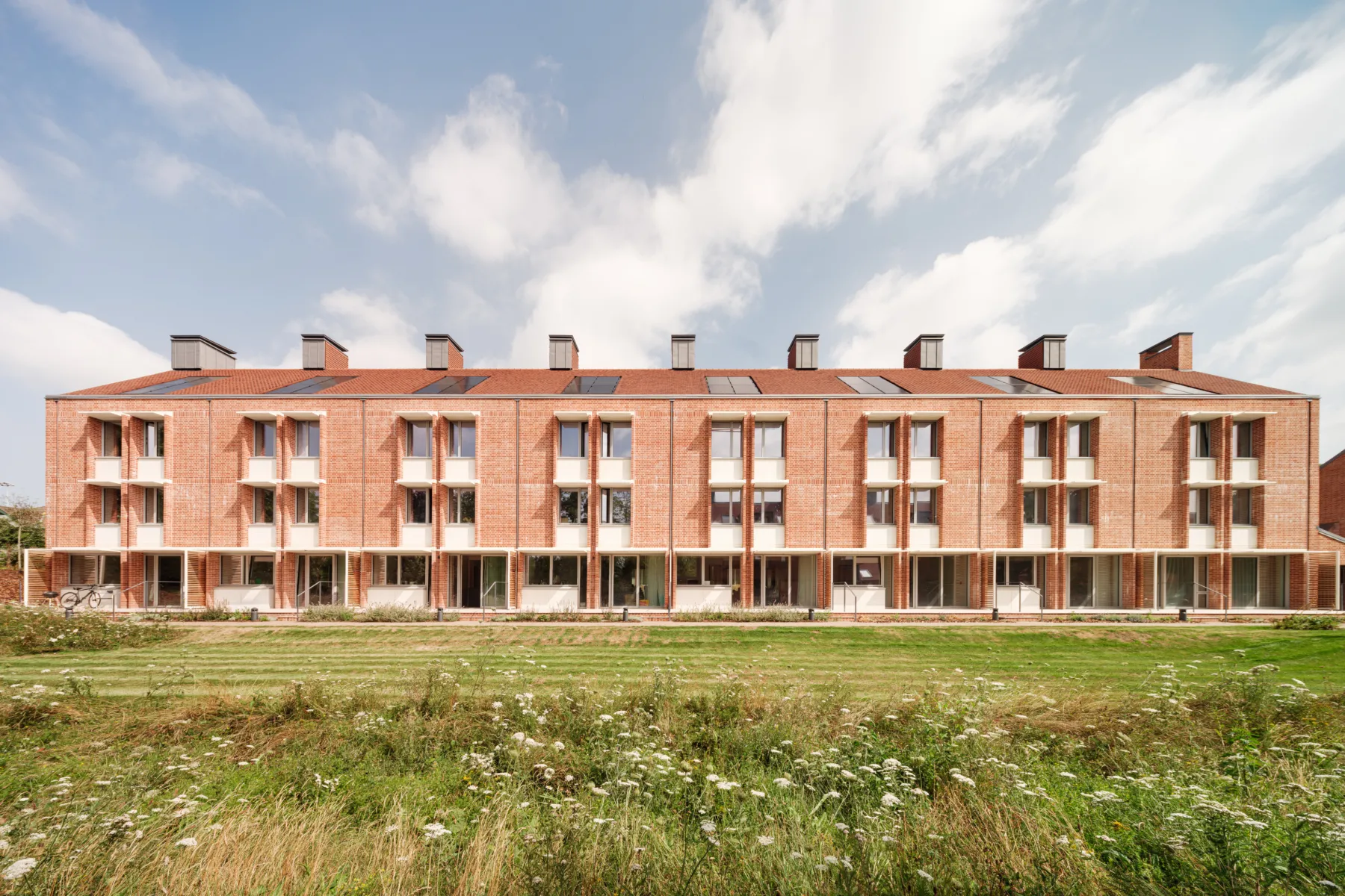 South-facing elevation of a red brick Passivhaus residential building with repetitive balconies and generous glazing, overlooking a meadow-like landscaped foreground under a partly cloudy sky.