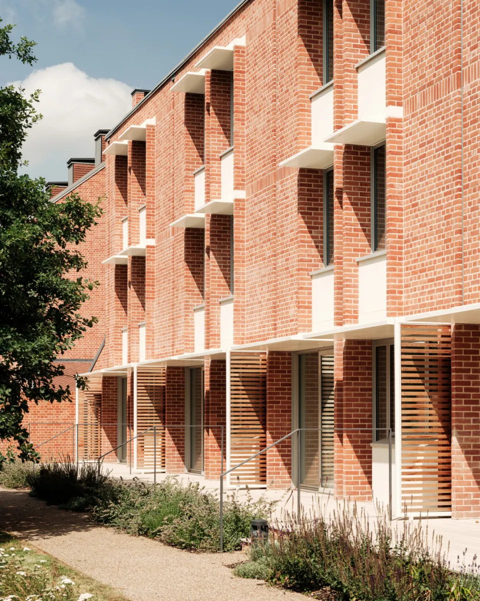 Close view of a red brick residential façade with repeating windows, small projecting balconies, and timber louvre screens at ground level, bordered by planting and a pedestrian path.