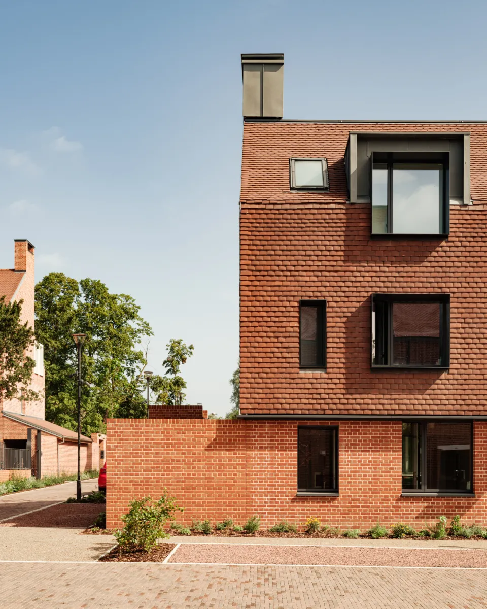 Corner view of a red brick residential building with pitched tiled roof, chimneys, and dormer windows. The building sits behind paved paths and planting, with trees and blue sky beyond.