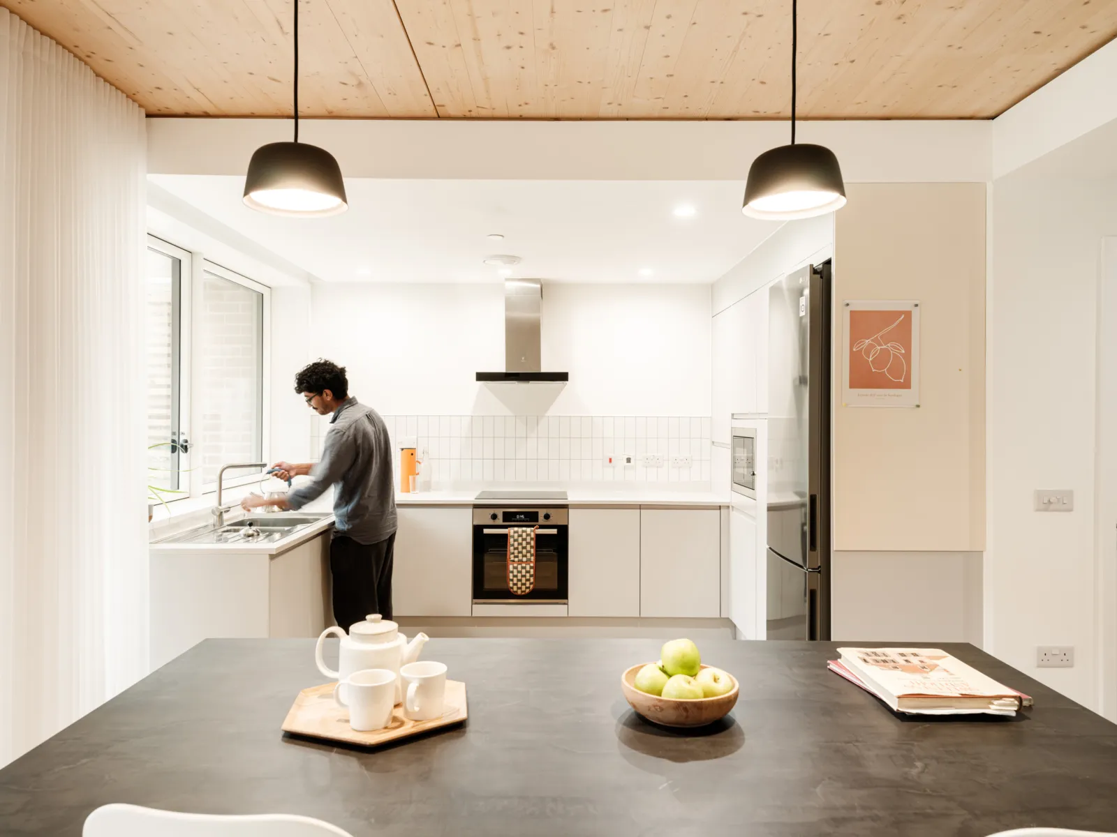 Open-plan shared kitchen with a timber ceiling and white cabinetry. A person stands at the sink while a central island with crockery and fruit sits in the foreground, lit by pendant lights and daylight from large windows.