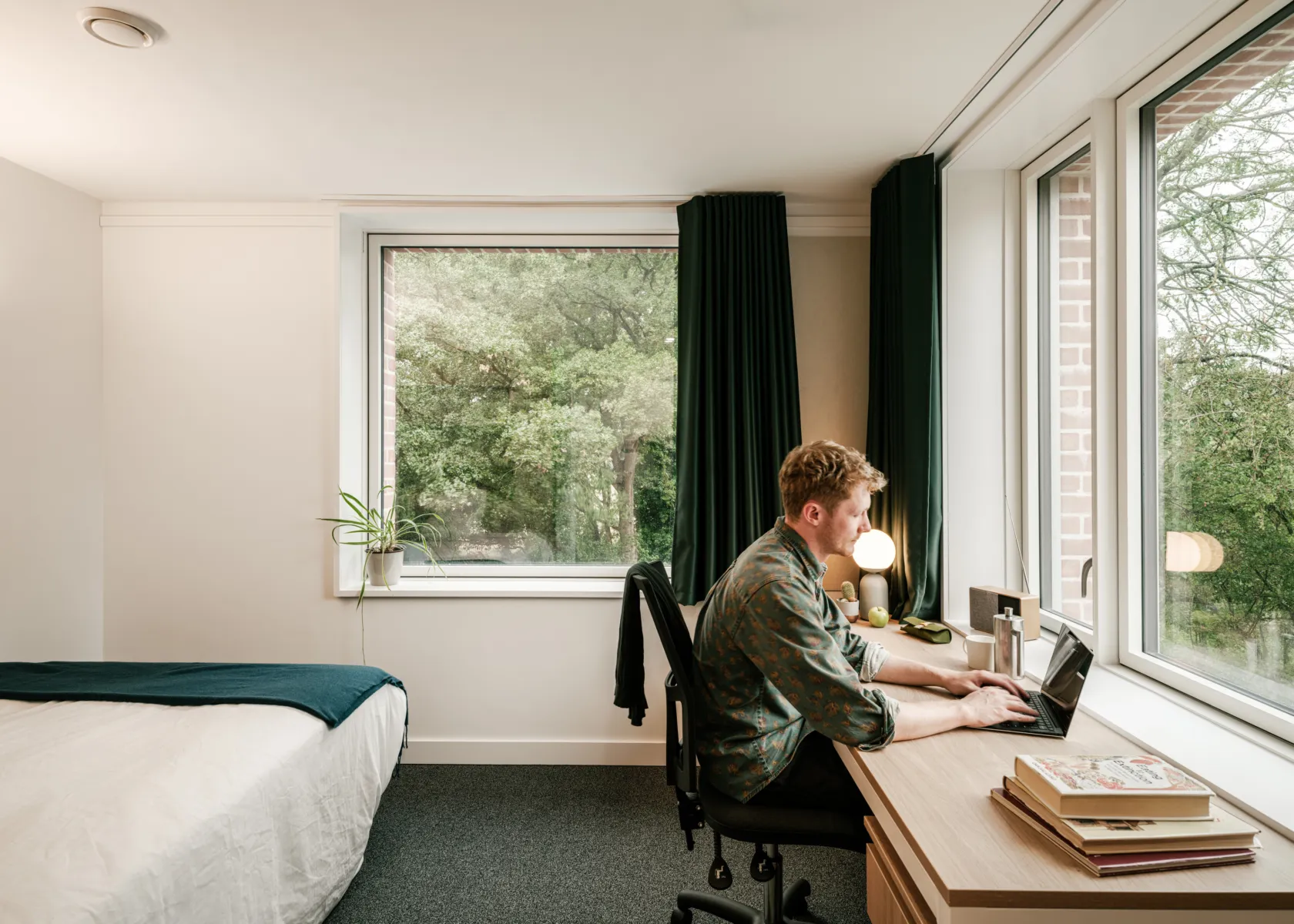 Student bedroom with a desk by a large window overlooking trees. A person sits working on a laptop, with a bed, desk lamp, books, and curtains visible in a bright, simply furnished space.