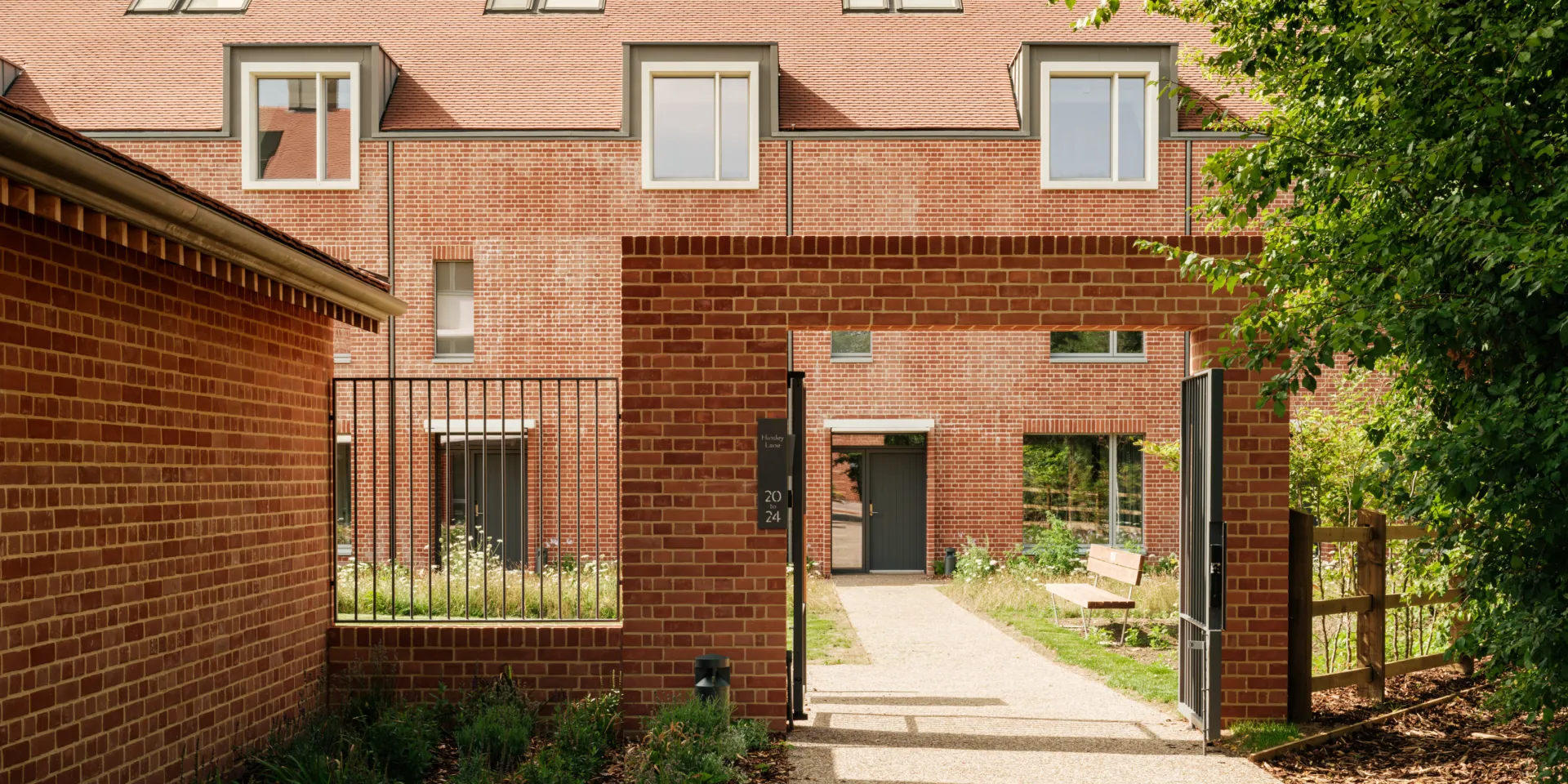 View through a brick gateway into a landscaped courtyard, with red brick terraces beyond featuring pitched tiled roofs, chimneys, and dormer windows. A gravel path leads to a residential entrance, with planting and trees framing the space.