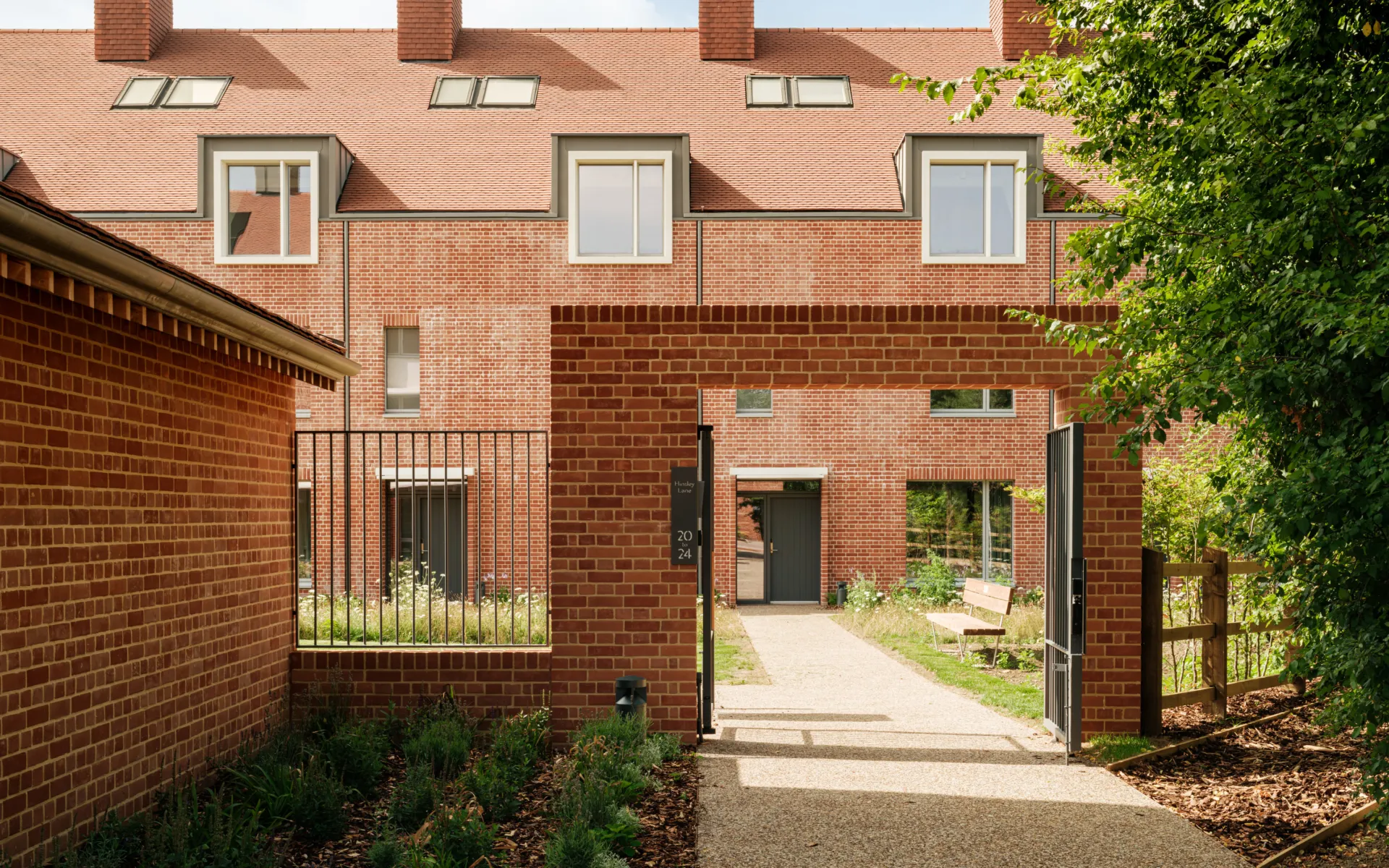 View through a brick gateway into a landscaped courtyard, with red brick terraces beyond featuring pitched tiled roofs, chimneys, and dormer windows. A gravel path leads to a residential entrance, with planting and trees framing the space.