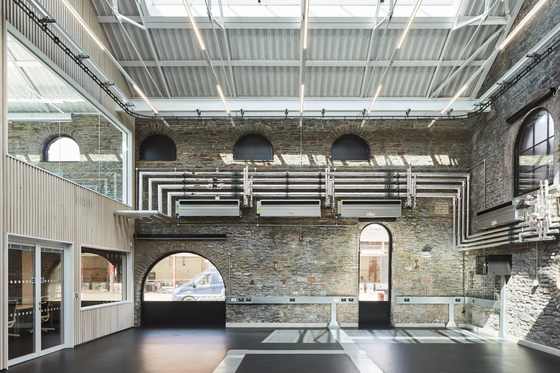 Images shows a brick wall with a series of pipes running across. The wall has two windows. The image also shows a ceiling with skylights and wooden wall with windows
