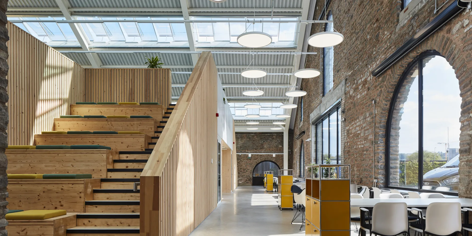image shows a large wooden hellerup stair case which allows for seating as well as stairs. The image shows a brick wall with large windows as well as and a ceiling with skylights.