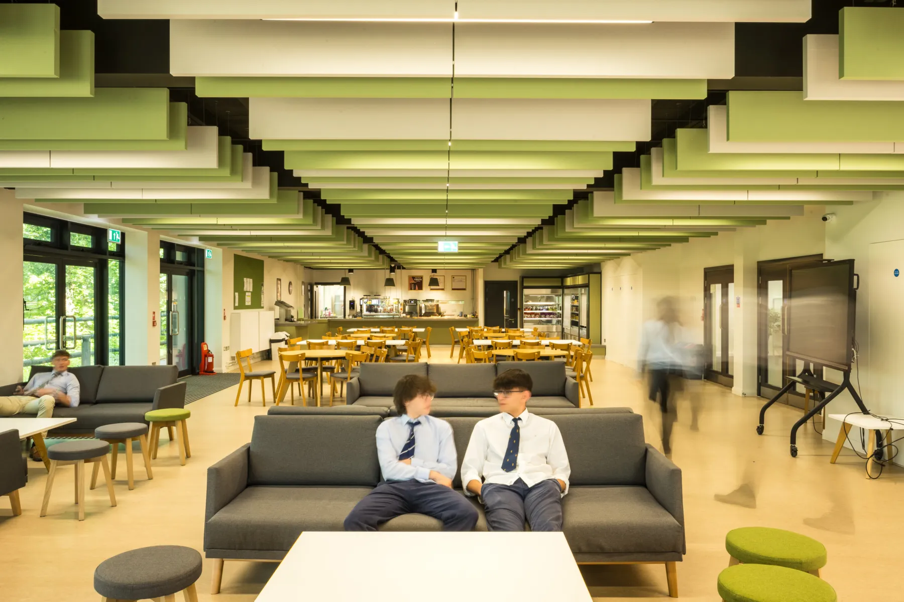 School dining and social space with green acoustic ceiling panels and students seated on sofas.