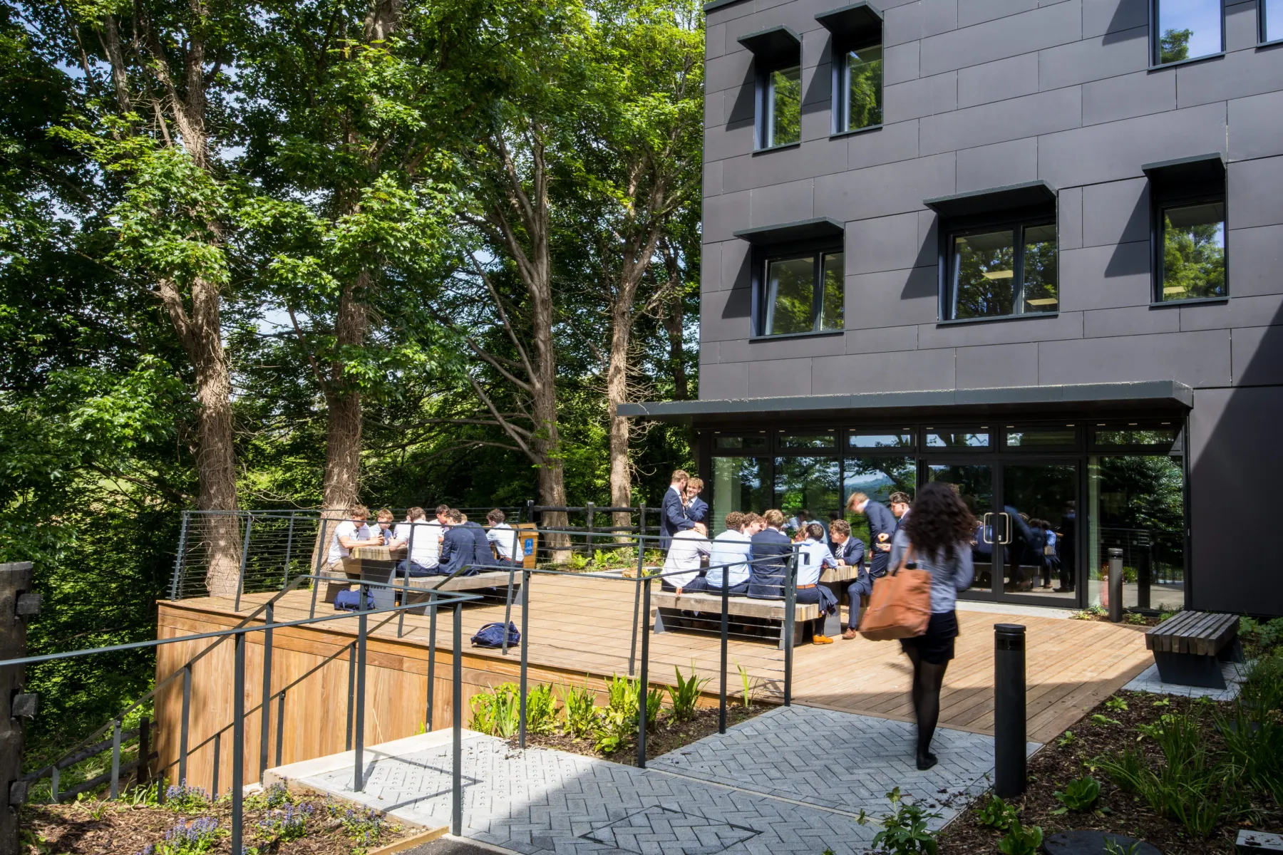 Outdoor terrace with students sitting at benches beside a modern school building and trees.