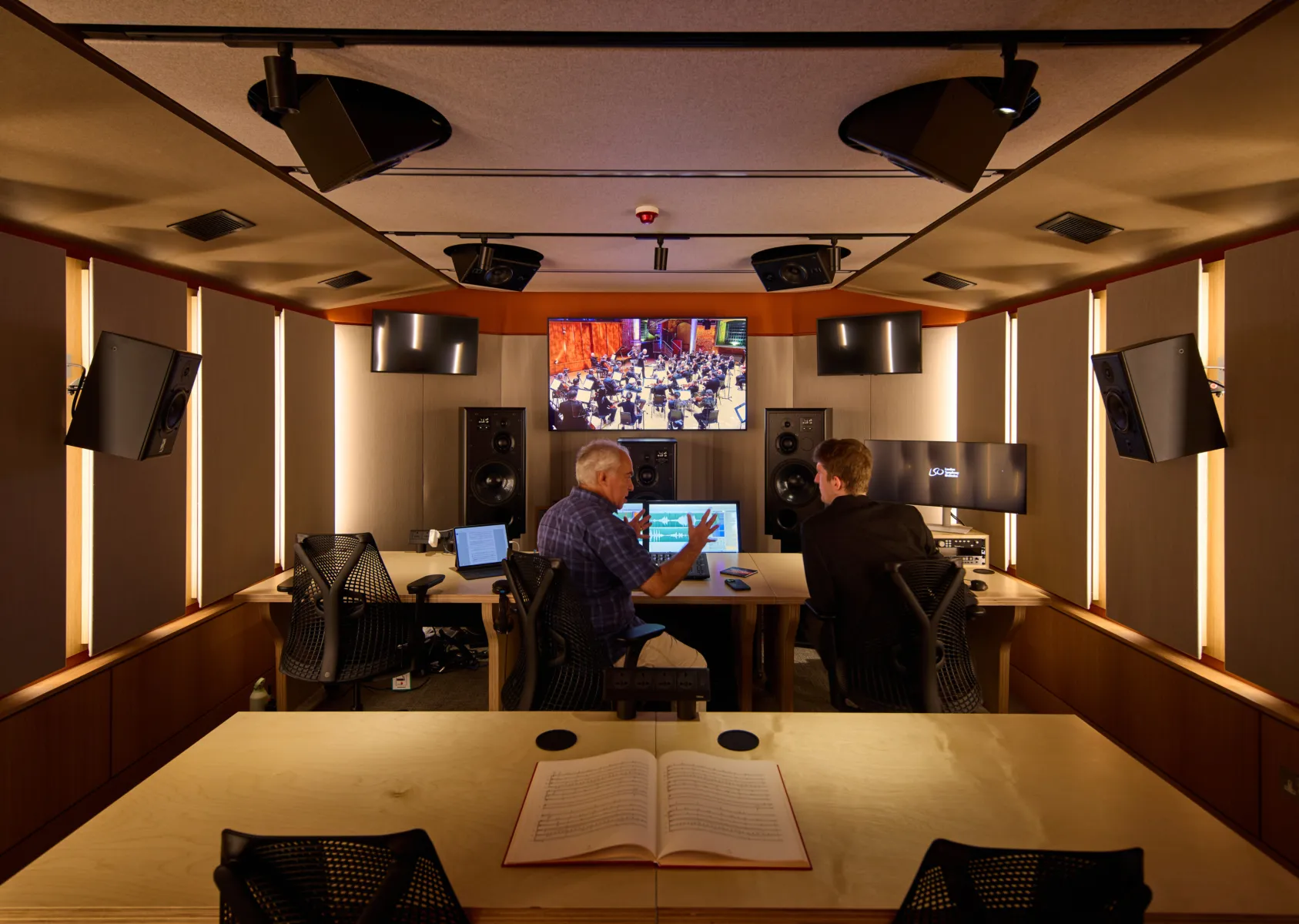 Two people in a sound control room reviewing audio with speakers and screens.