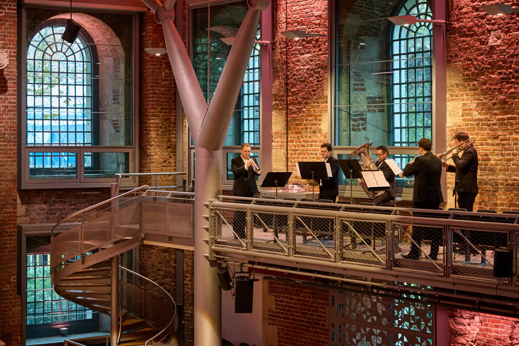 Brass ensemble performing on a balcony inside a brick-lined auditorium with tall windows.