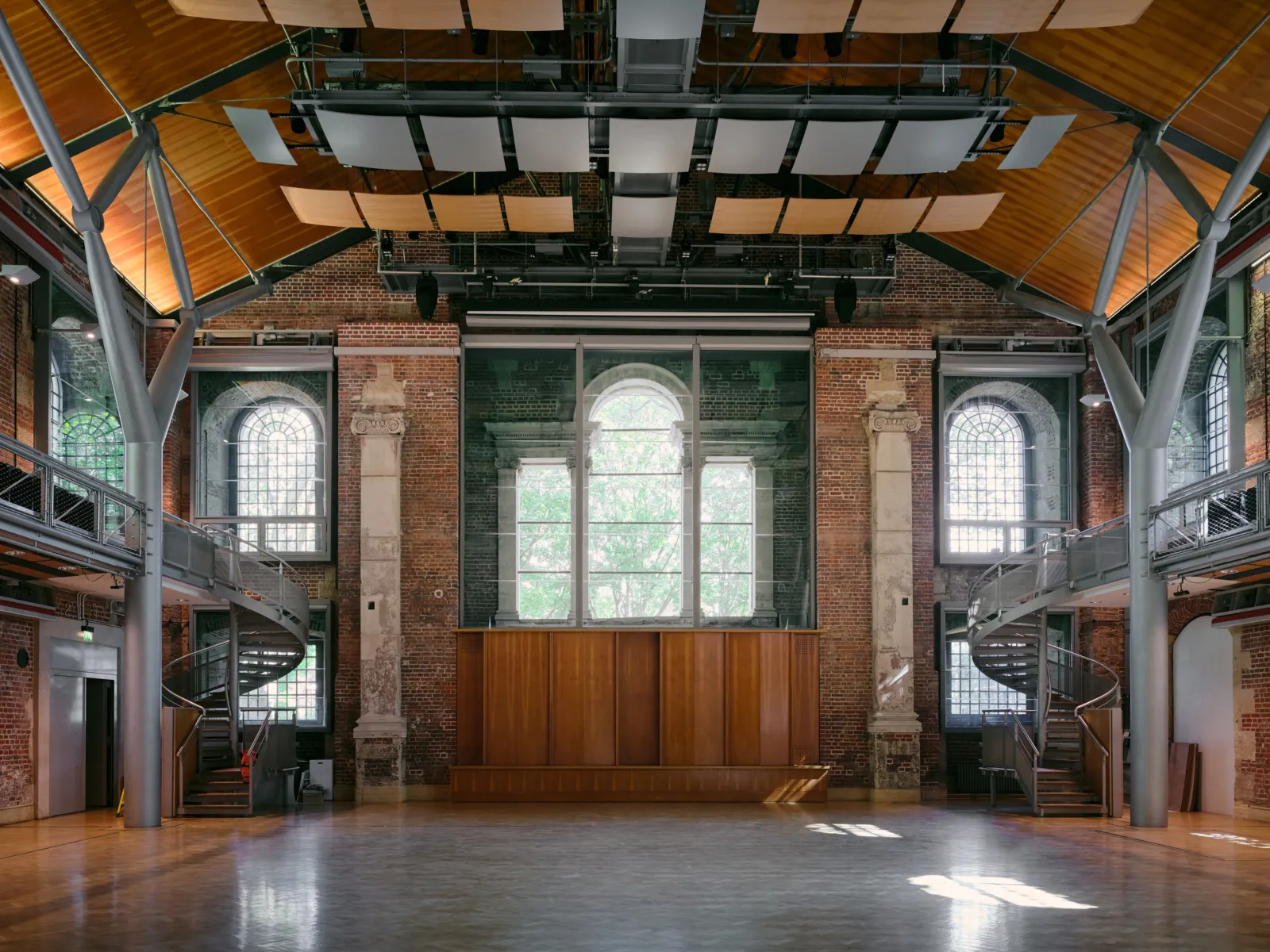 Empty auditorium with brick walls, large windows, balconies, and overhead acoustic panels.