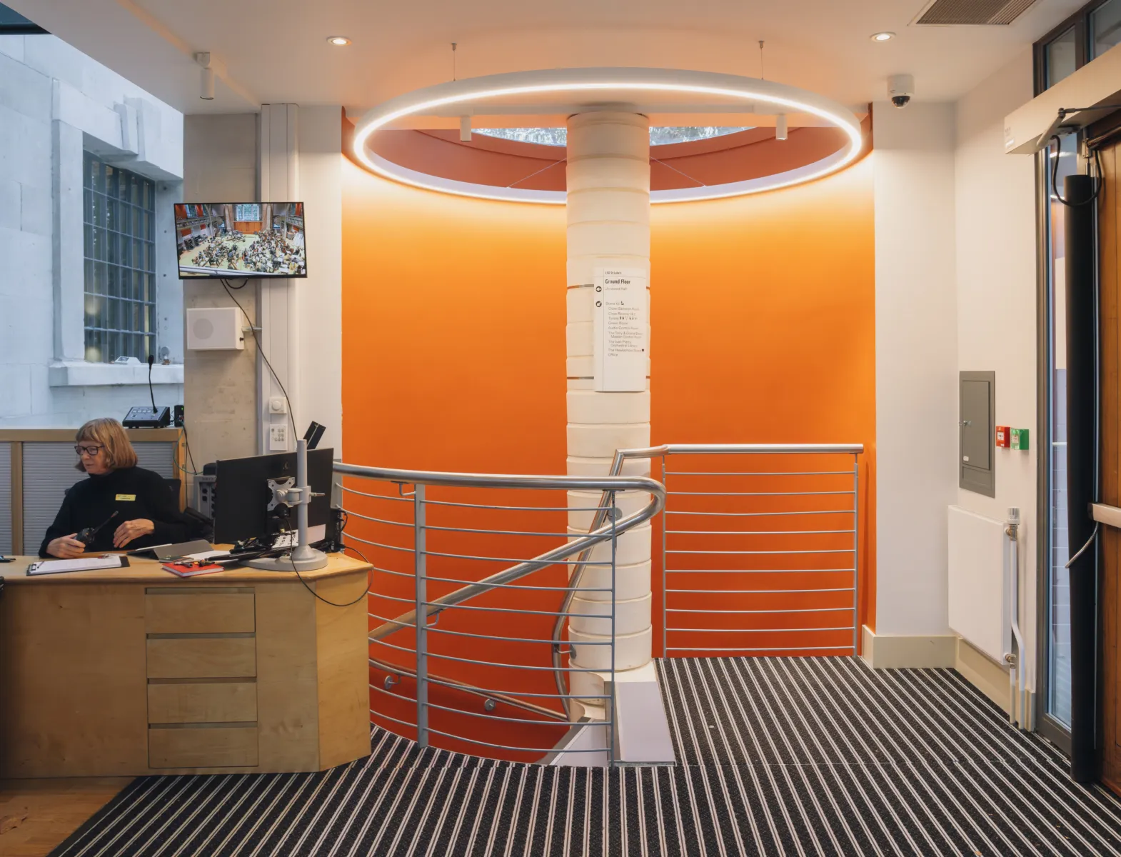 Reception desk beside an orange wall and stair with circular ceiling light above.