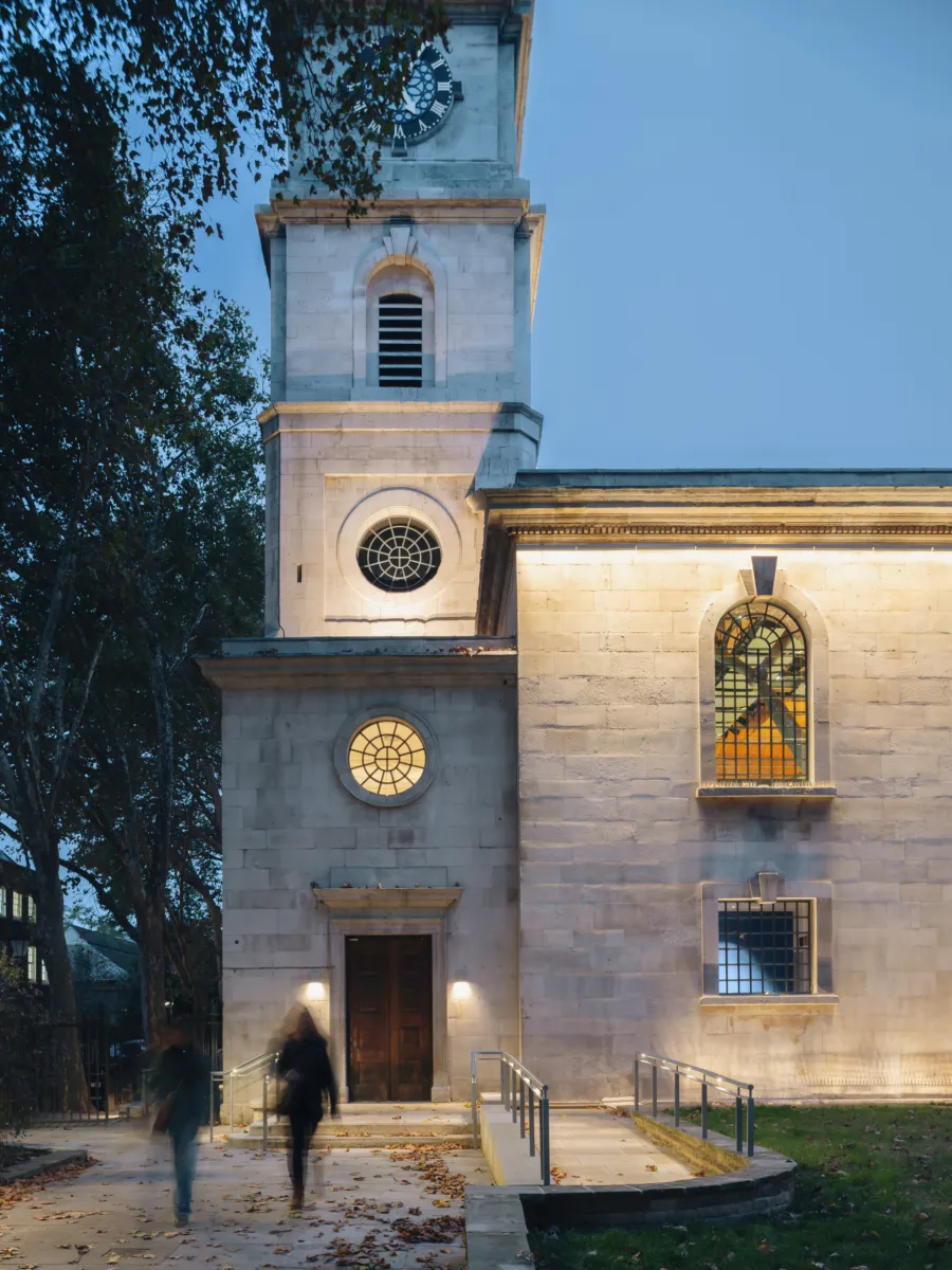 Close-up of building façade with tower clock, arched windows, and lit entrance at dusk.