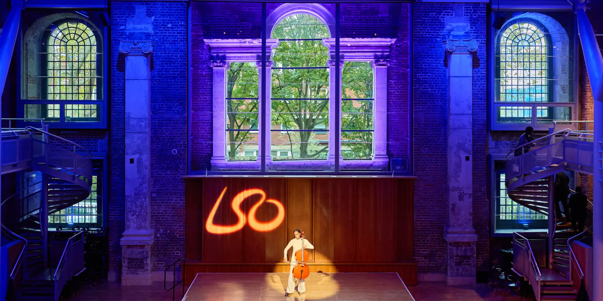 Cellist performing solo on a stage under coloured lighting in a historic hall.