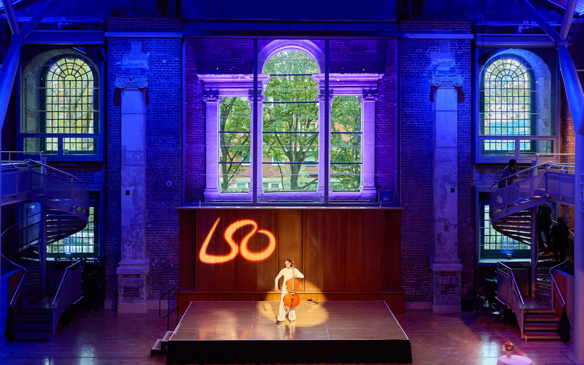 Cellist performing solo on a stage under coloured lighting in a historic hall.