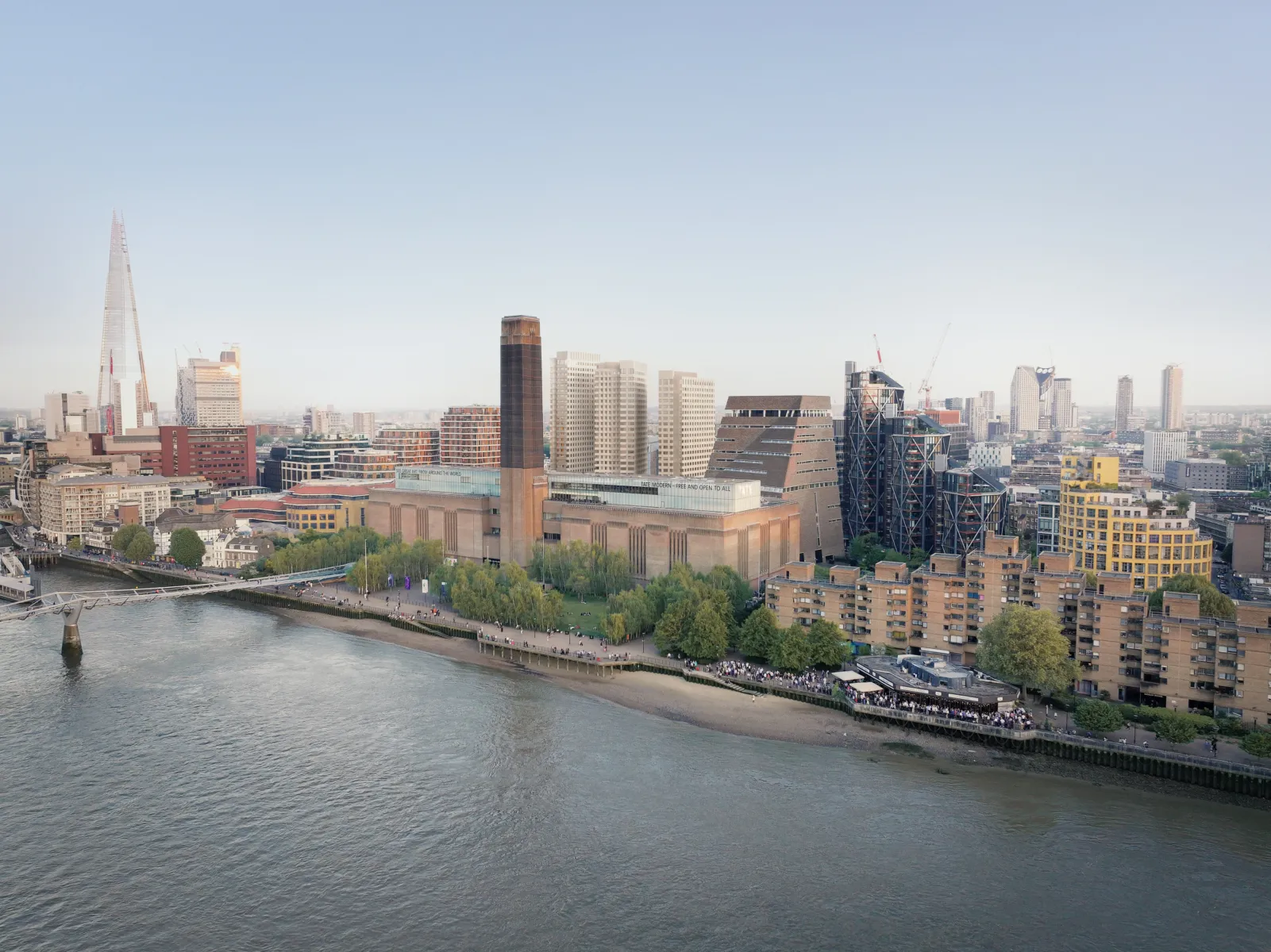 A wide aerial view across the River Thames showing Tate Modern with its central brick chimney, the Millennium Bridge at left, and the London skyline behind, including The Shard, under a clear sky.