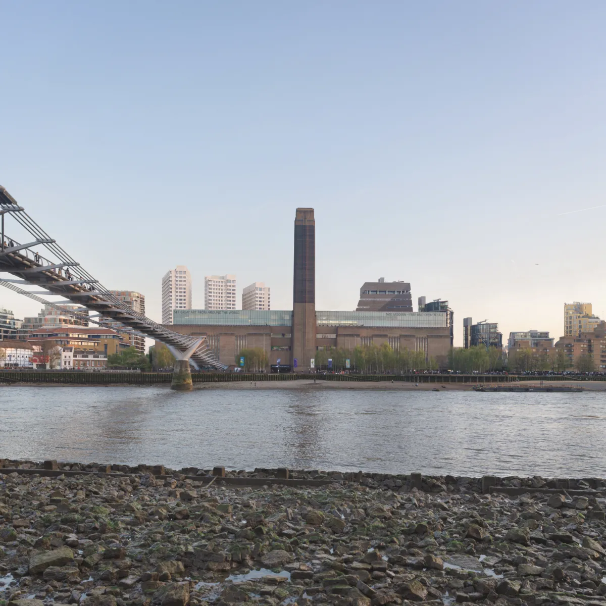 Tate Modern on the opposite bank of the River Thames, with the Millennium Bridge entering from the left and the gallery’s tall brick chimney centred, and a rocky shoreline in the foreground.
