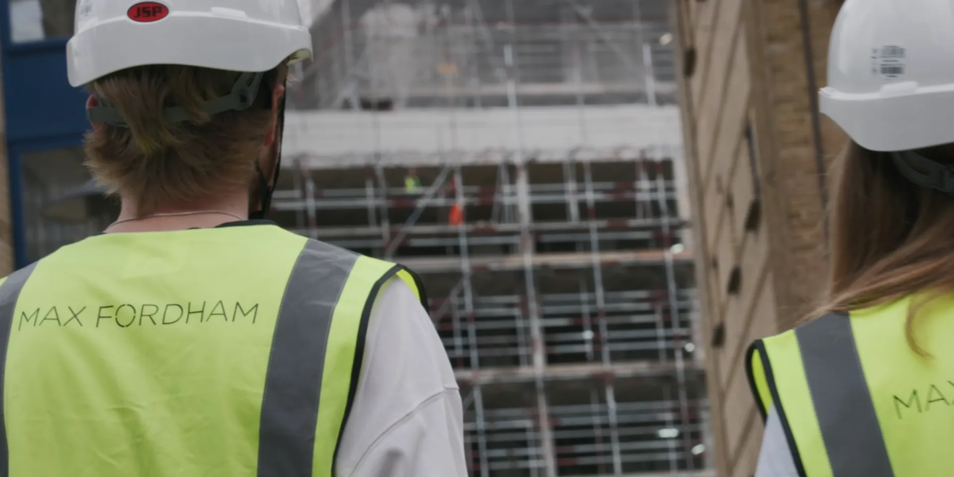 Two people wearing MAX FORDHAM site vests and hard hats looking at a building site