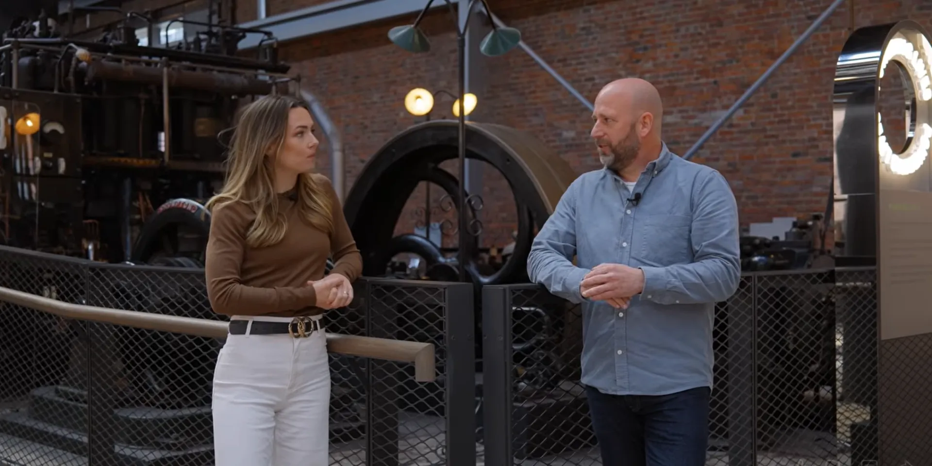 A man and a woman are stood on front of an old engine at a museum talking