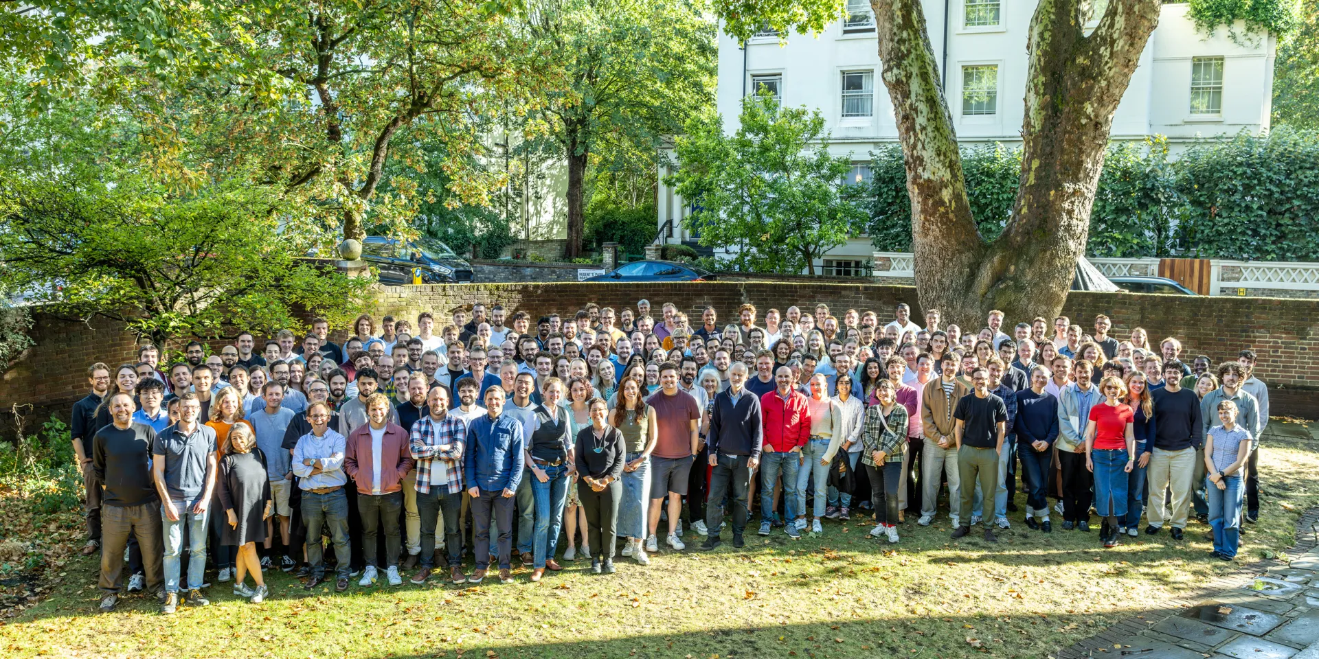 Large group of staff standing together on a lawn beneath mature trees for an outdoor team photograph.