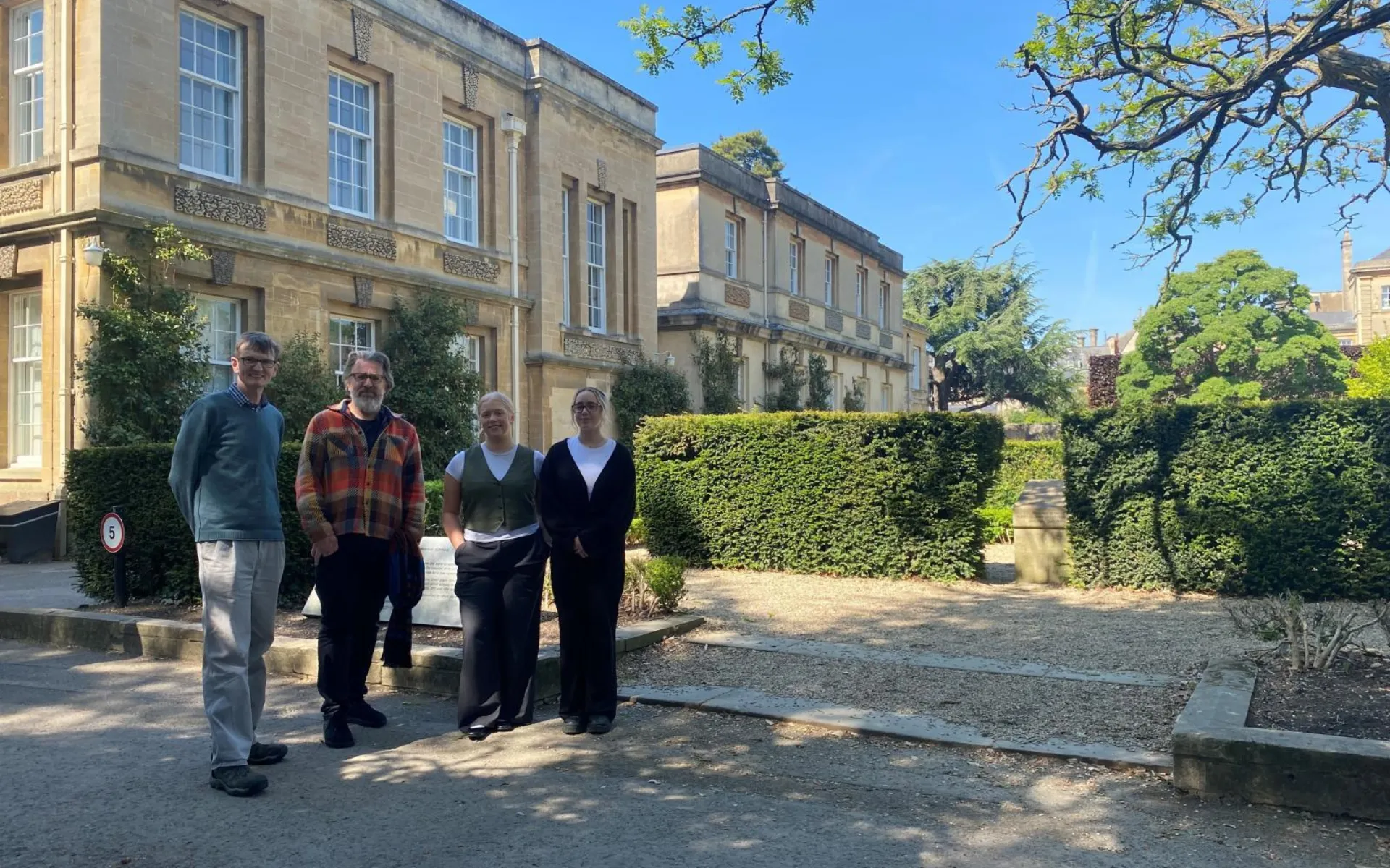 Two men and two women stand in front of a sandstone historic building at Magdalen College, Oxford