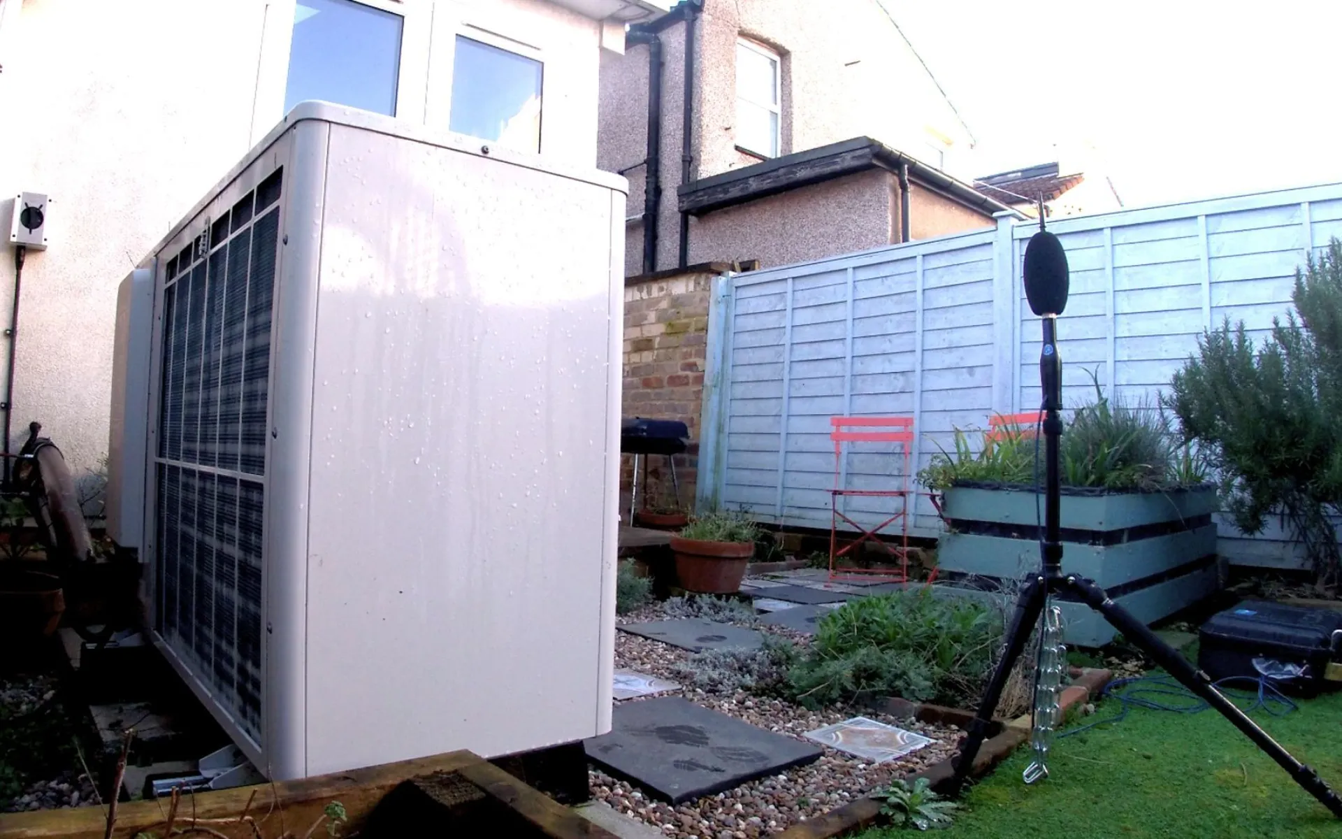 Images show a large, white rectangular box, which is an air source heat pump, in the garden next to a house. There is recording equipment positioned to record the sound coming from the air source heat pump
