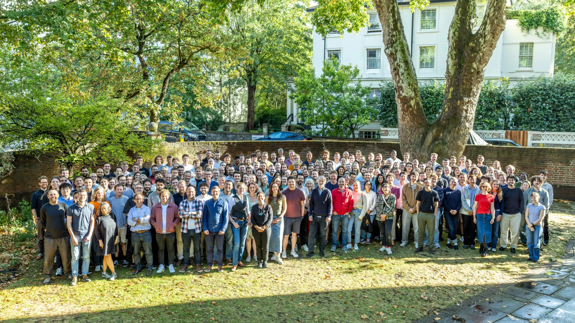 Large group of staff standing together on a lawn beneath mature trees for an outdoor team photograph.