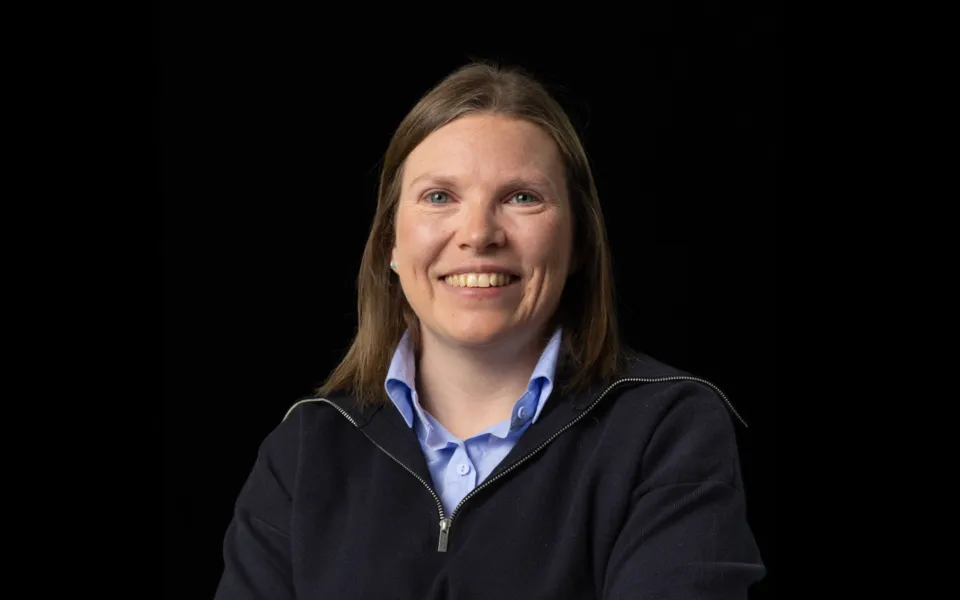 Portrait of Kathryn Donald wearing a light blue shirt under a dark zipped jumper, against a black background.